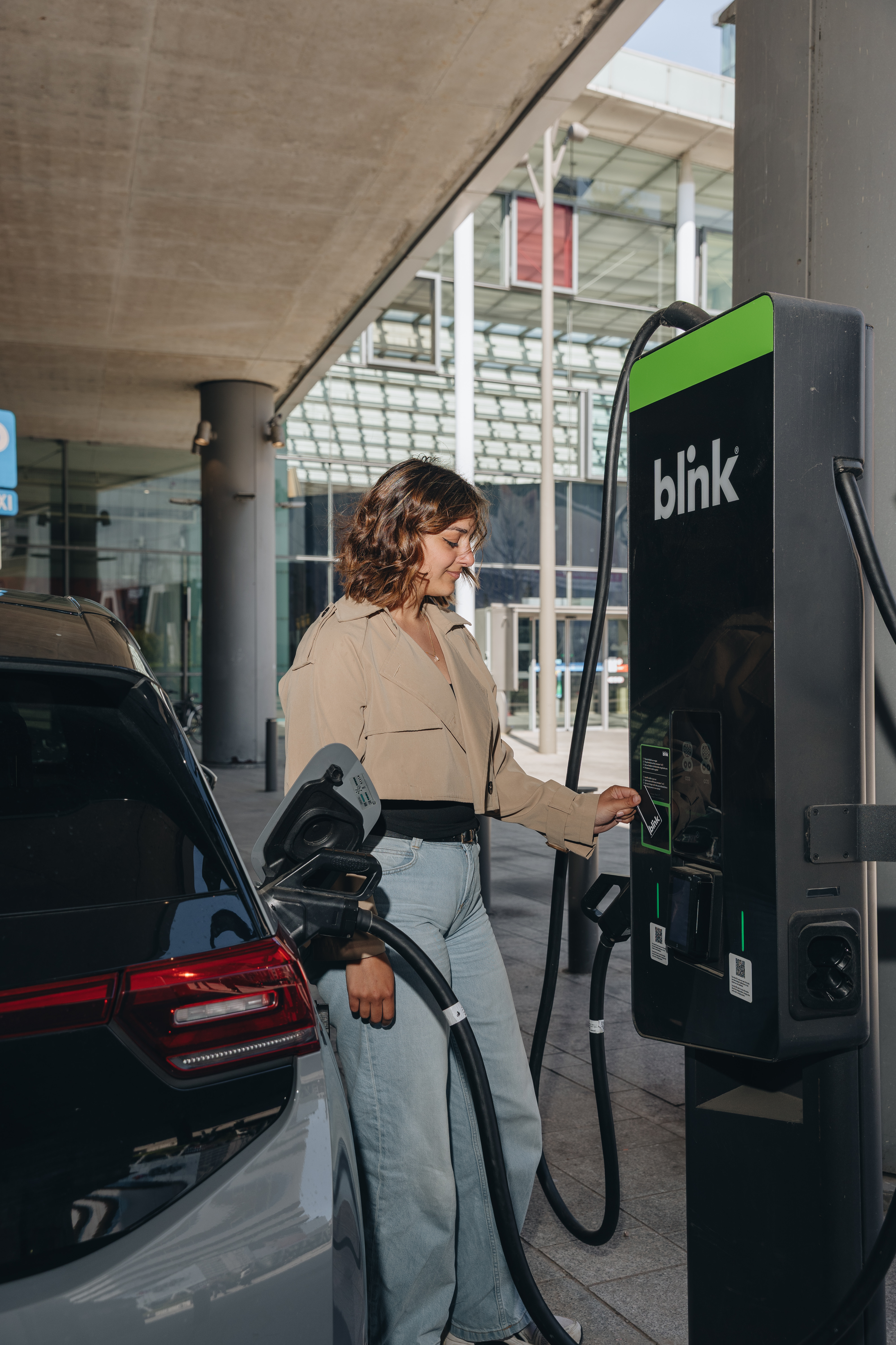 Woman charging an electric car at a Blink station, under a modern building with glass details.