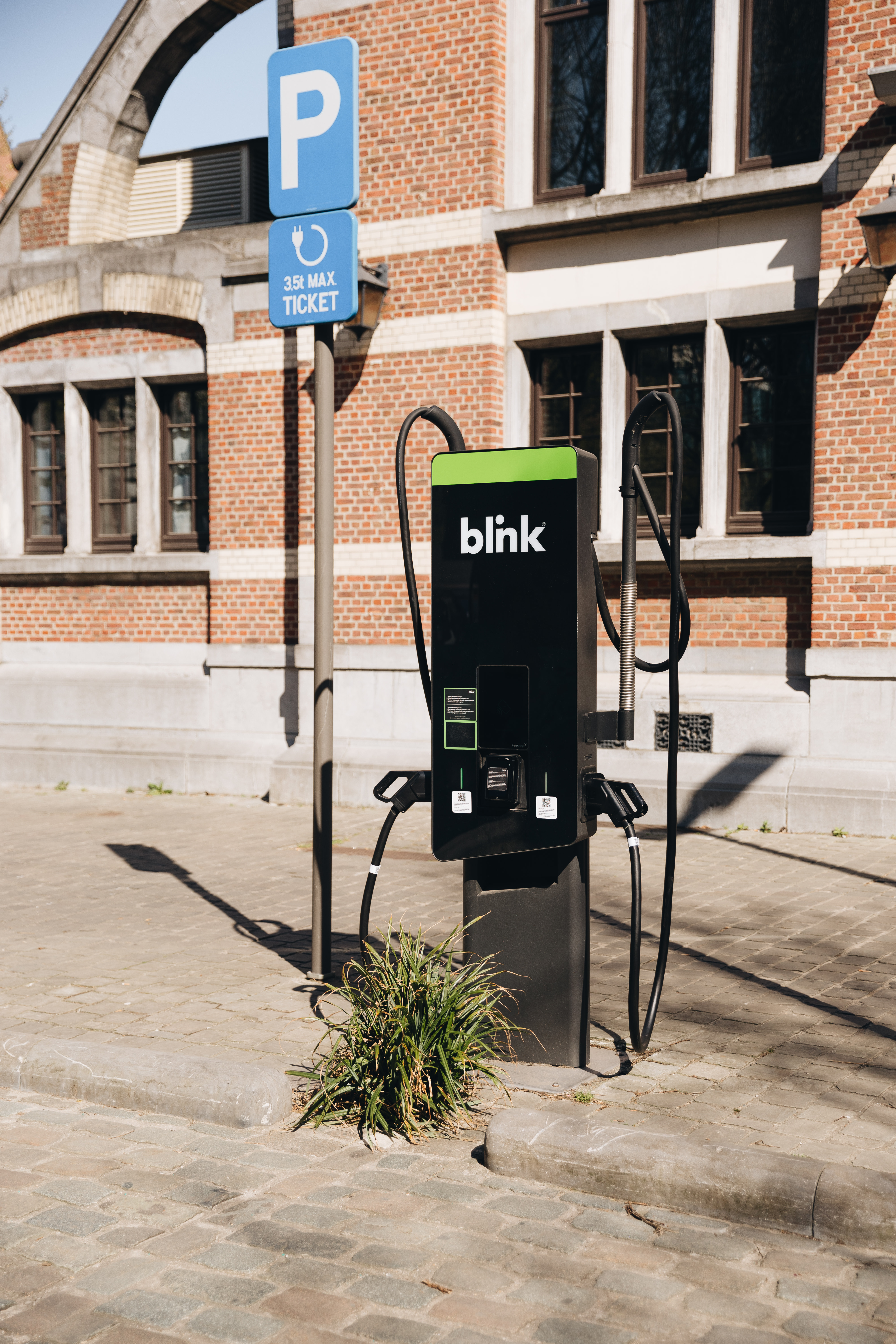 Electric vehicle charging station with "blink" logo in urban setting, near brick building and parking sign.
