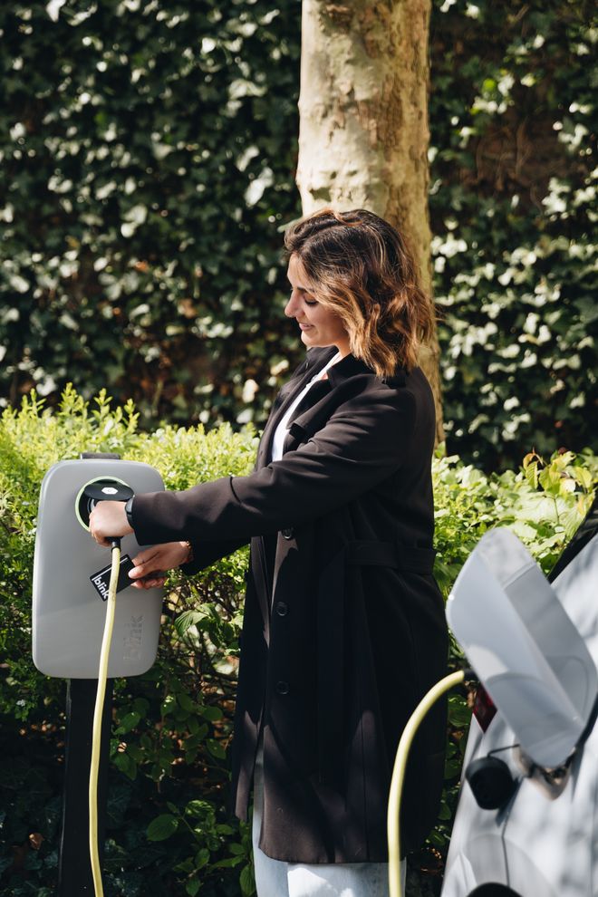 A woman in a black coat connects a charging cable to an electric car at a charging station, surrounded by greenery.
