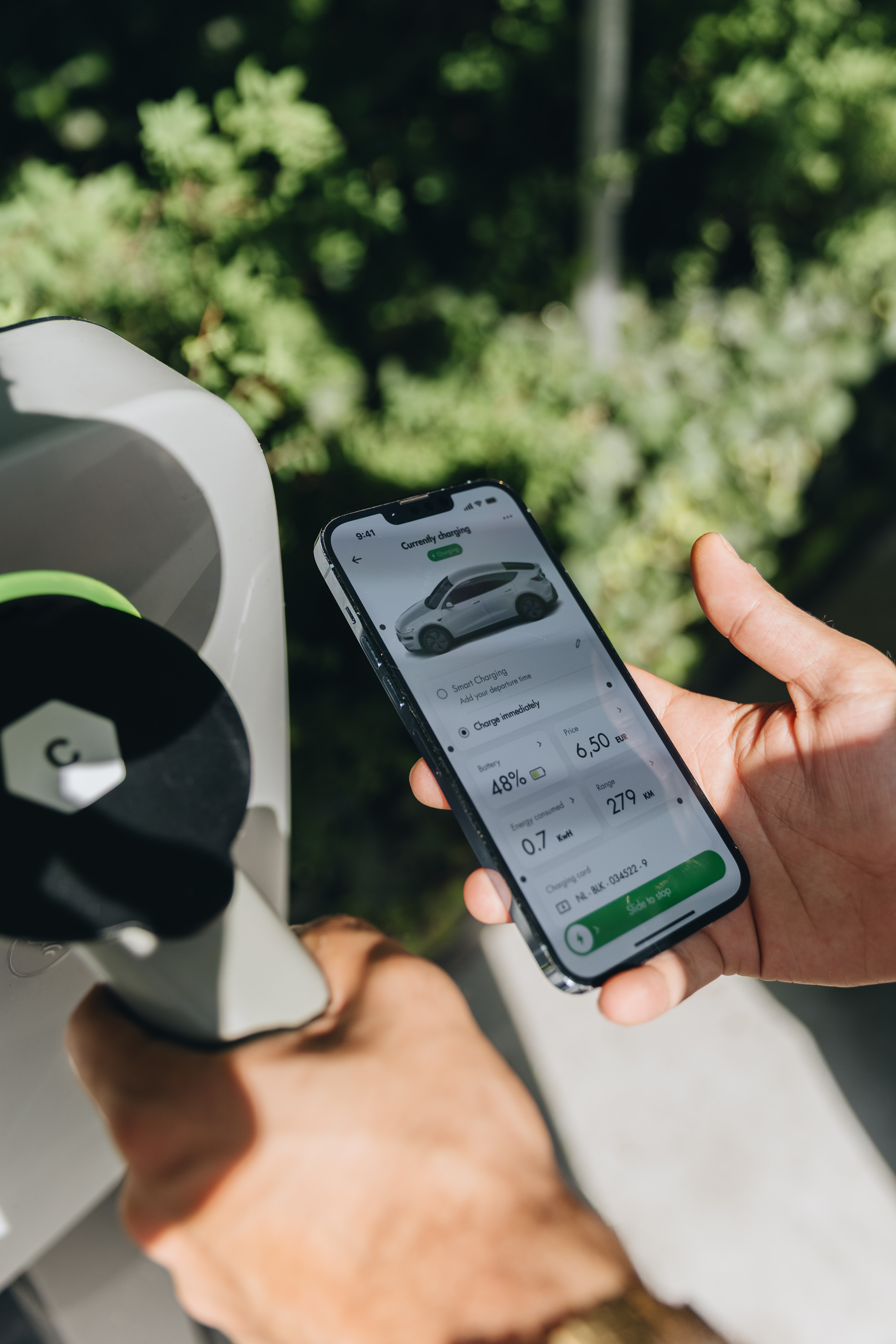 Person holding a smartphone displaying EV charging status and a charging plug, set against a leafy background.