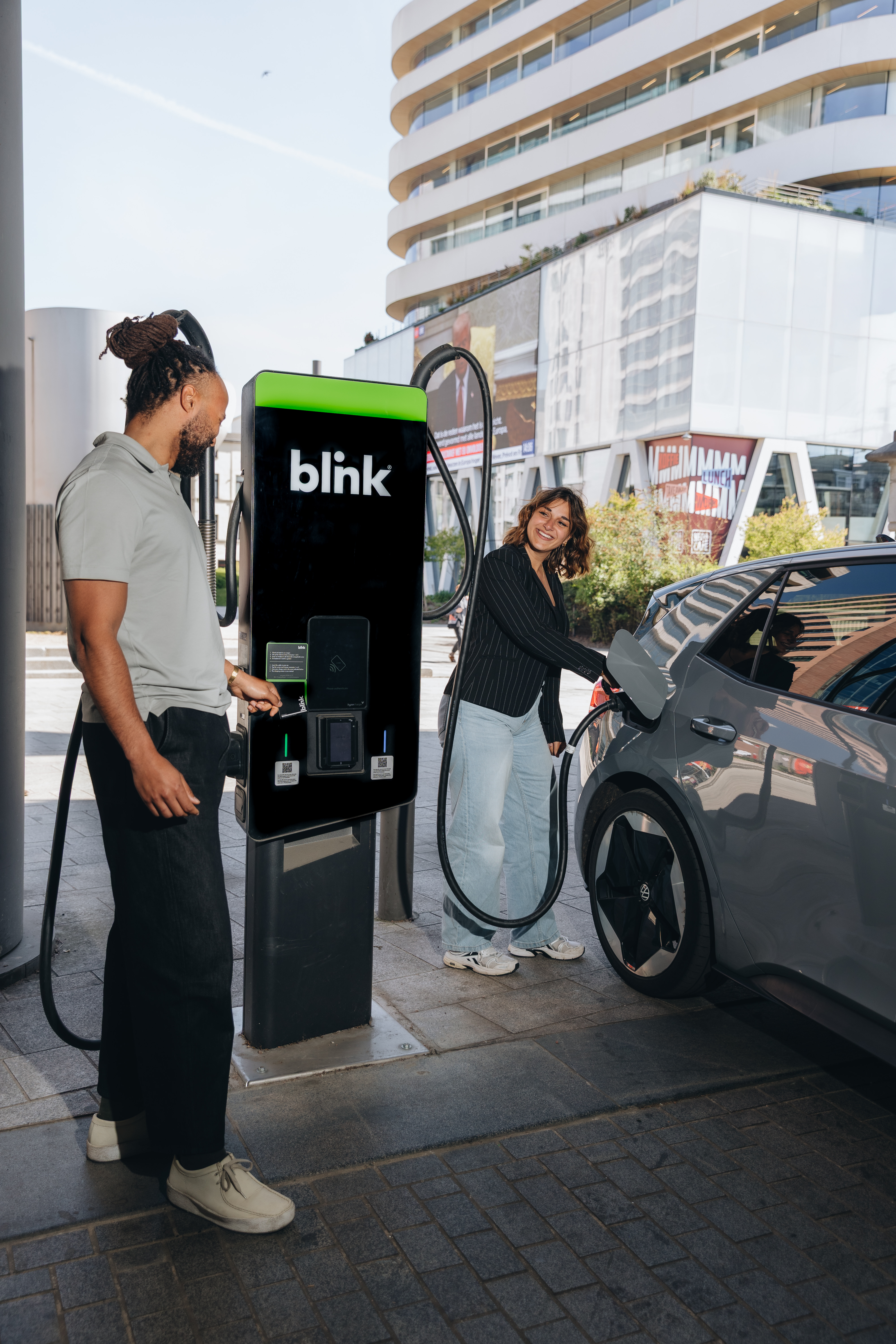 Two people charging an electric car at a Blink charging station outdoors, in a modern urban setting.