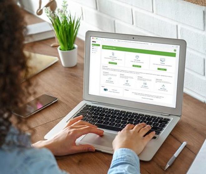 Person typing on a laptop displaying a webpage, next to a smartphone and a potted plant on a wooden desk.