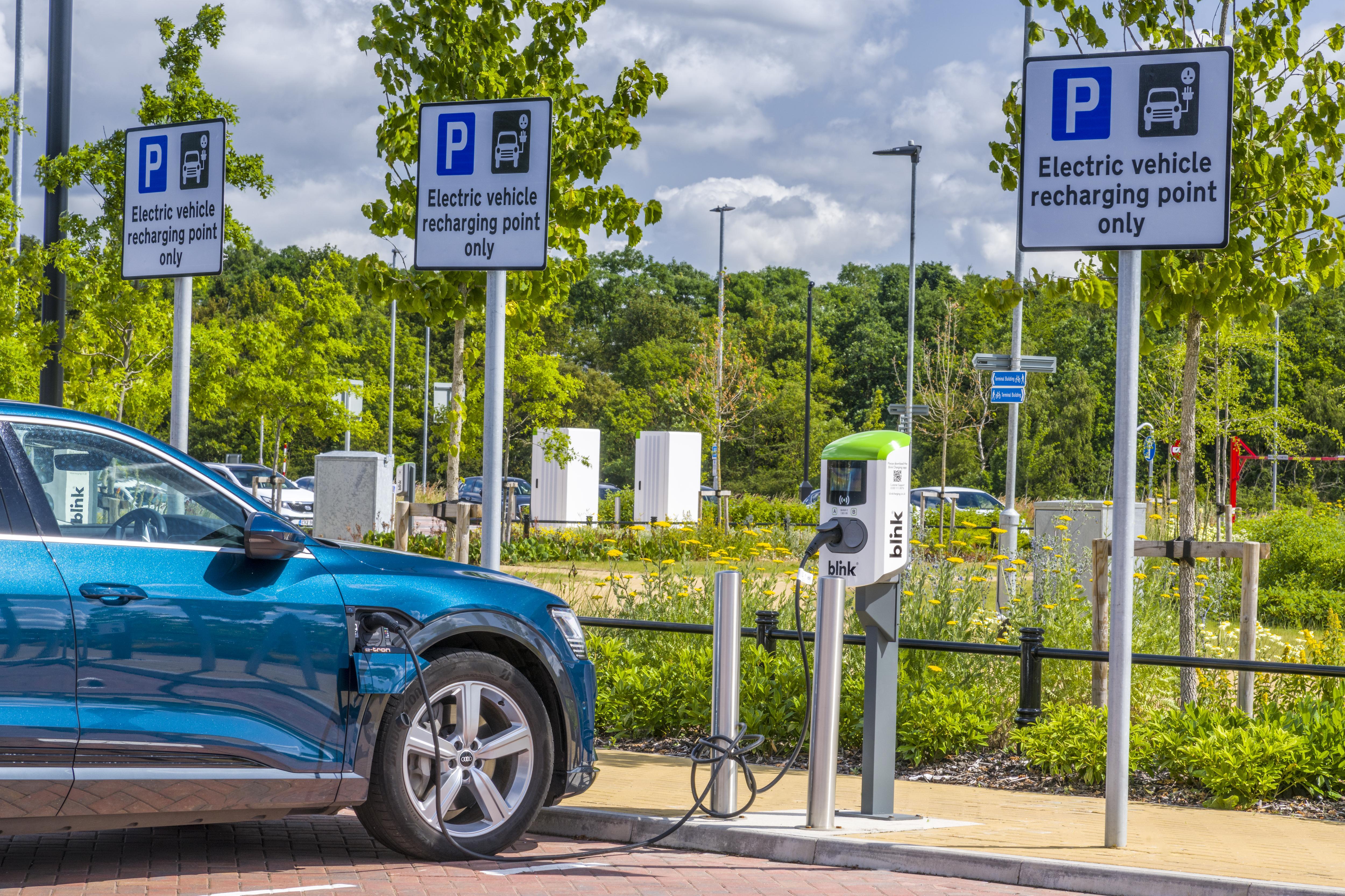 Electric car charging at a station in a parking lot with signs reading "Electric vehicle recharging point only," surrounded by trees and greenery.