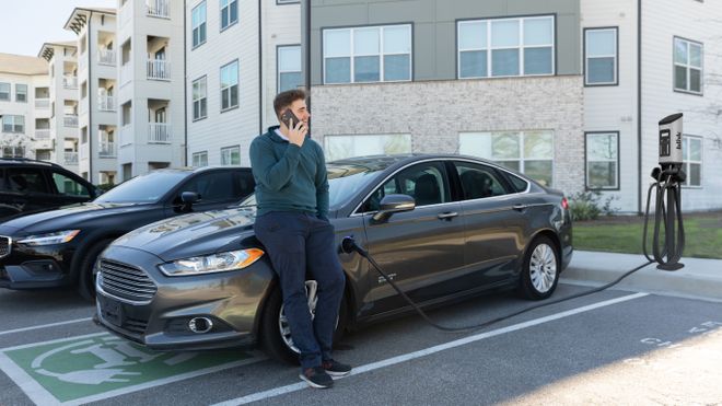 Man talking on phone while leaning on an electric car being charged in a parking lot near modern apartments.