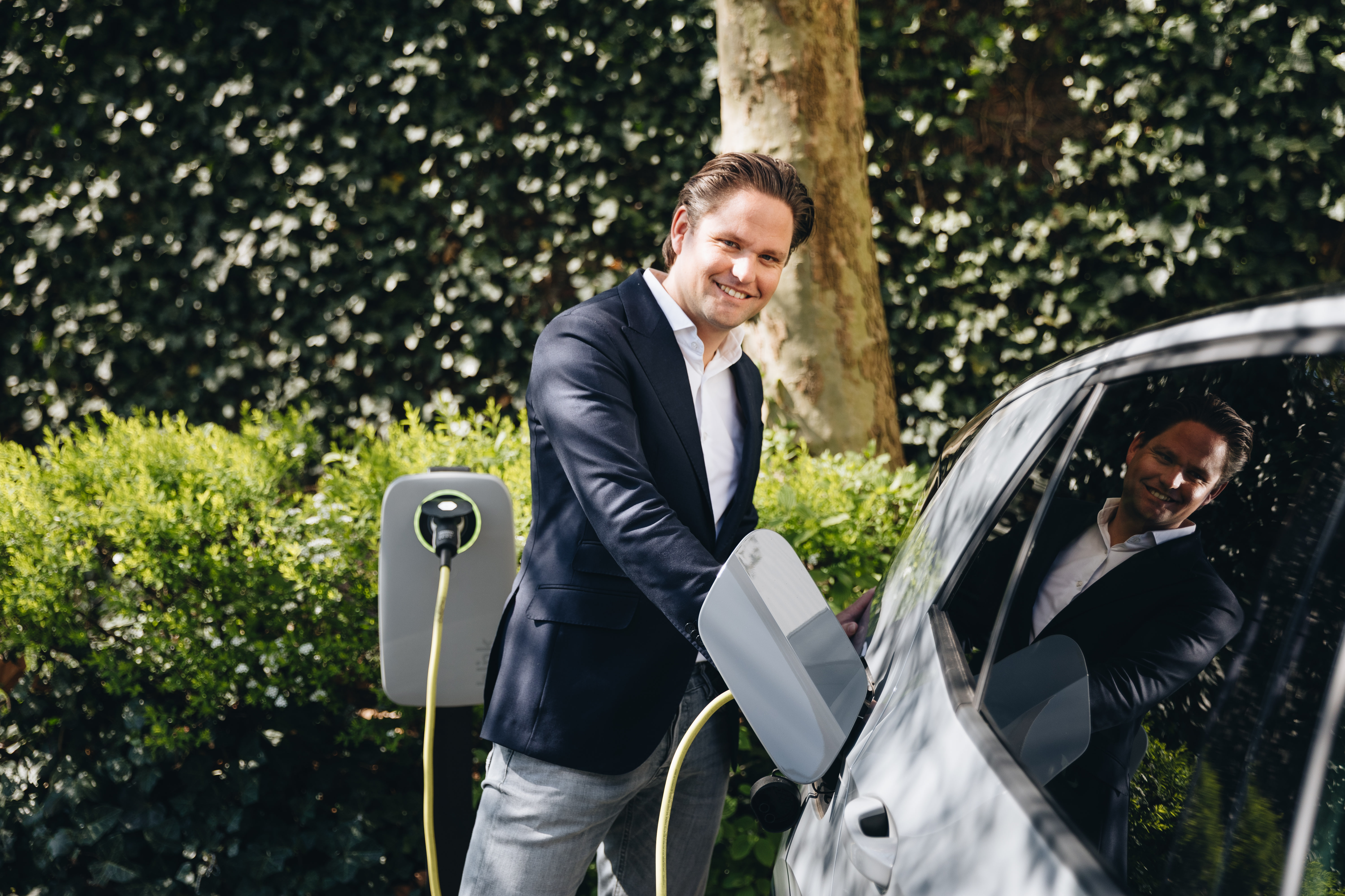 Man in a suit charging an electric car outdoors, standing by greenery with a smile, reflecting in the car window.