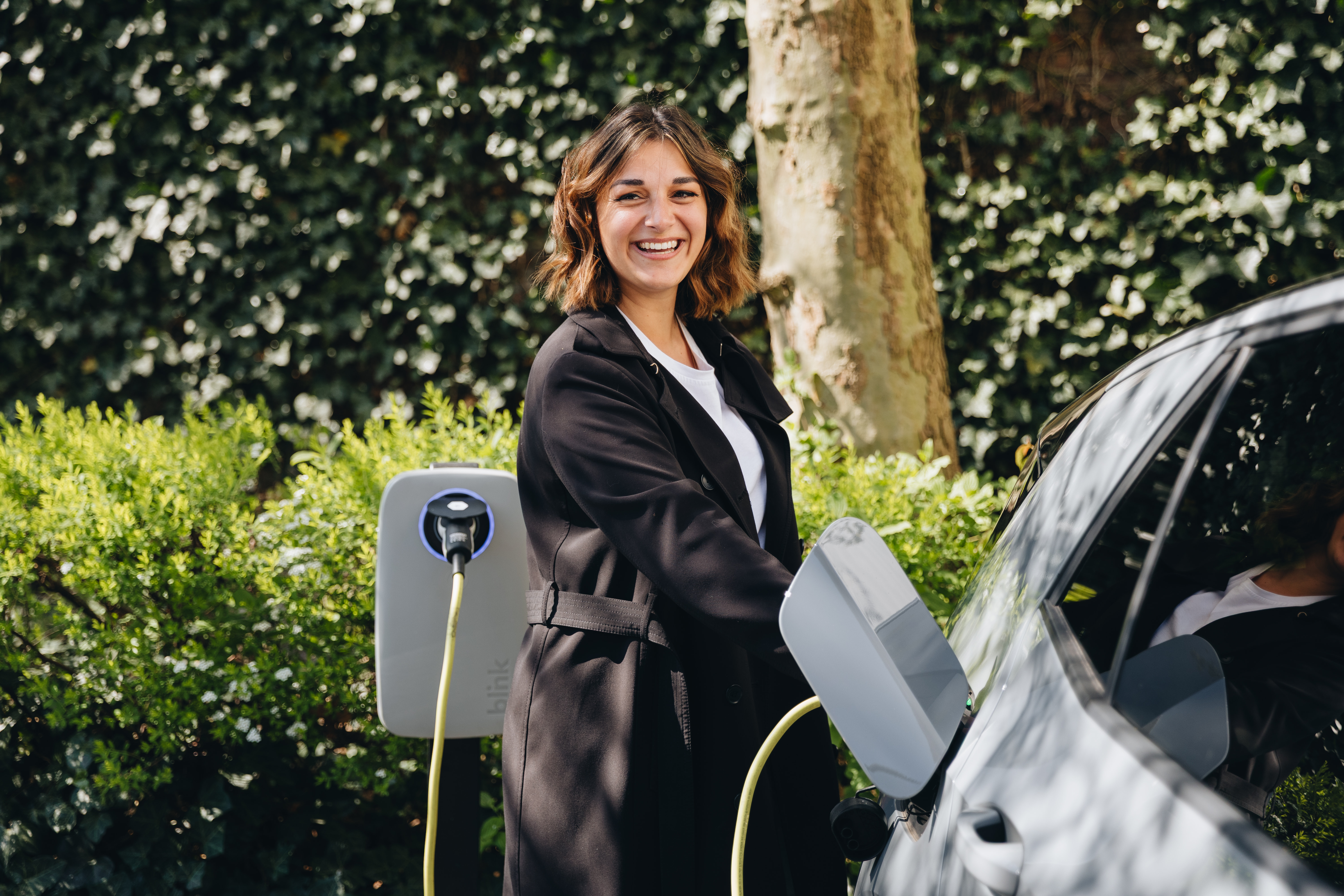 A woman smiling while charging an electric car at a charging station in a leafy outdoor setting.