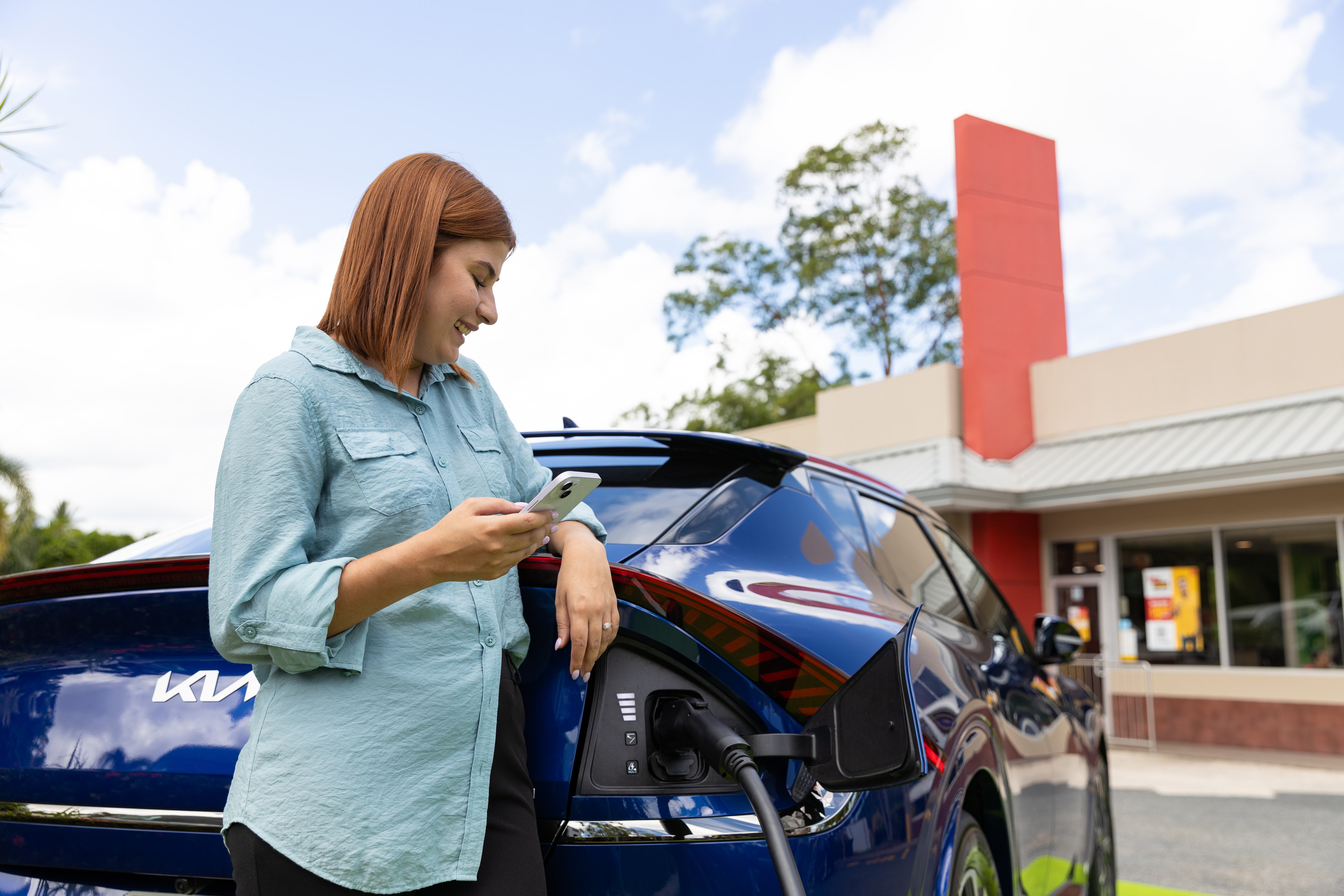 Woman in a blue shirt using a smartphone, leaning on a parked blue electric car that's charging, with a store in the background.