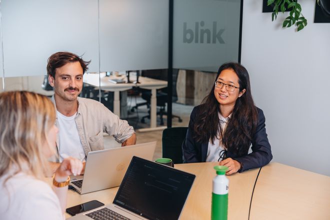 Three people seated around a table with laptops, engaged in a discussion inside a modern office with "blink" on a glass partition.
