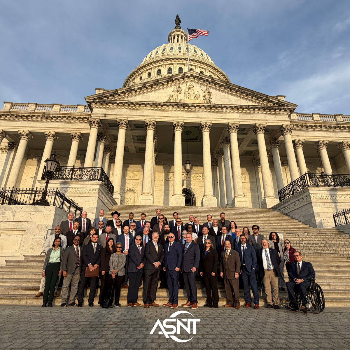 A large group of people in formal attire stand on the steps of the U.S. Capitol, with an American flag flying above. ASNT logo is visible.