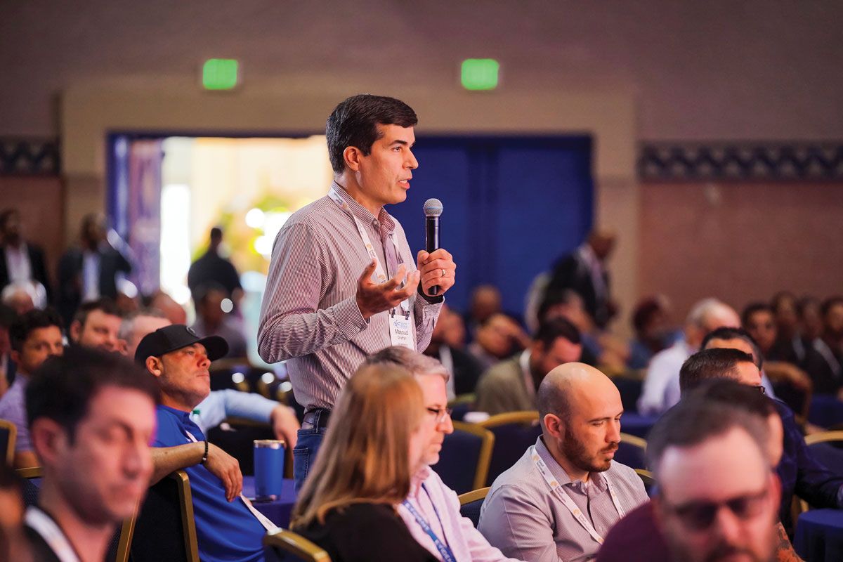 Photograph of an attendee standing with a microphone to ask a question during a Q&A session at a past ASNT conference, with other attendees seated nearby.