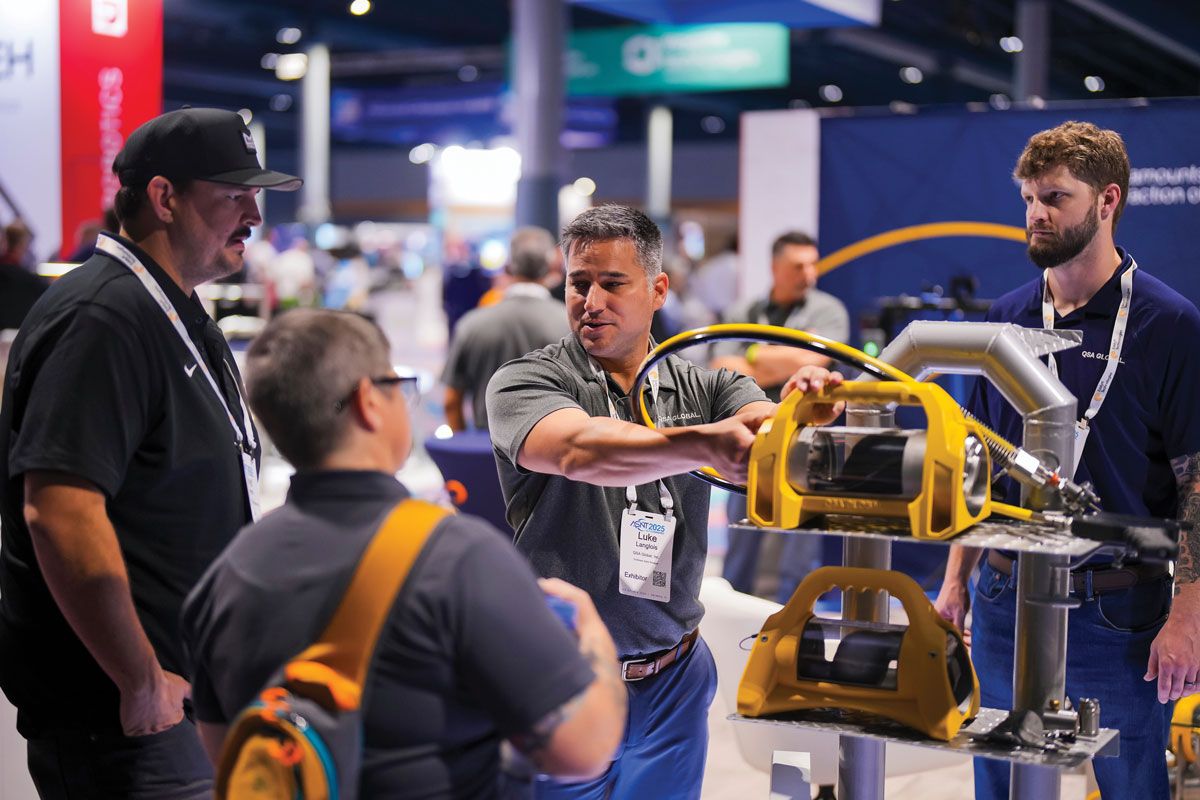 Photograph of the exhibit hall at a past ASNT conference, where an exhibitor demonstrates a yellow magnetic-particle testing yoke to a group of attendees.
