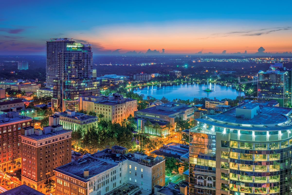 Aerial view of a cityscape at sunset with illuminated buildings, a central lake, and a colorful sky transitioning from orange to blue.