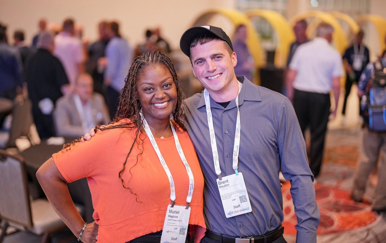 Two people smiling at a conference, wearing name tags and casual attire, with a blurred crowd in the background.
