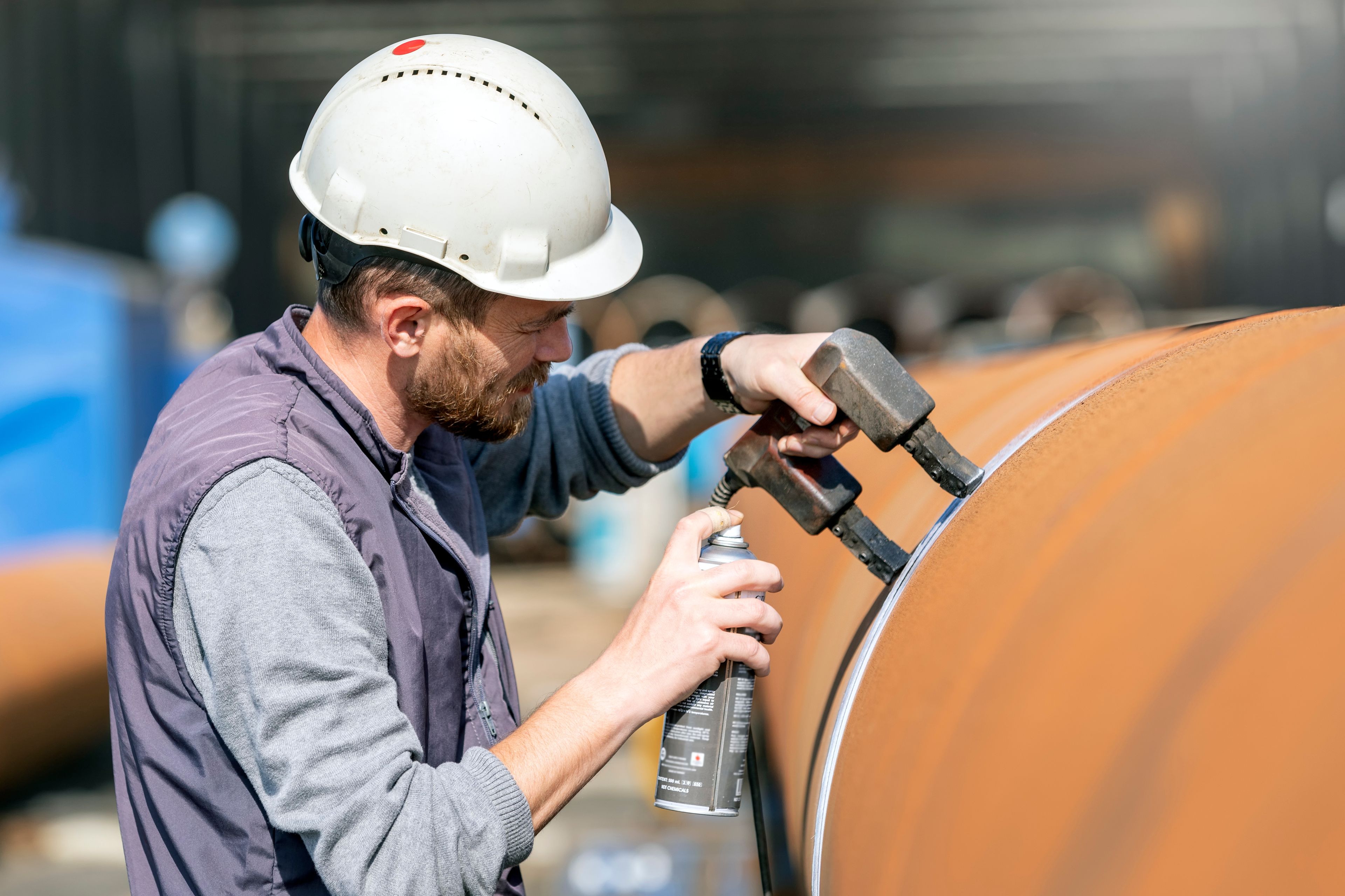 Technician in a hard hat performs magnetic particle inspection with a yoke and aerosol on a large steel pipe.