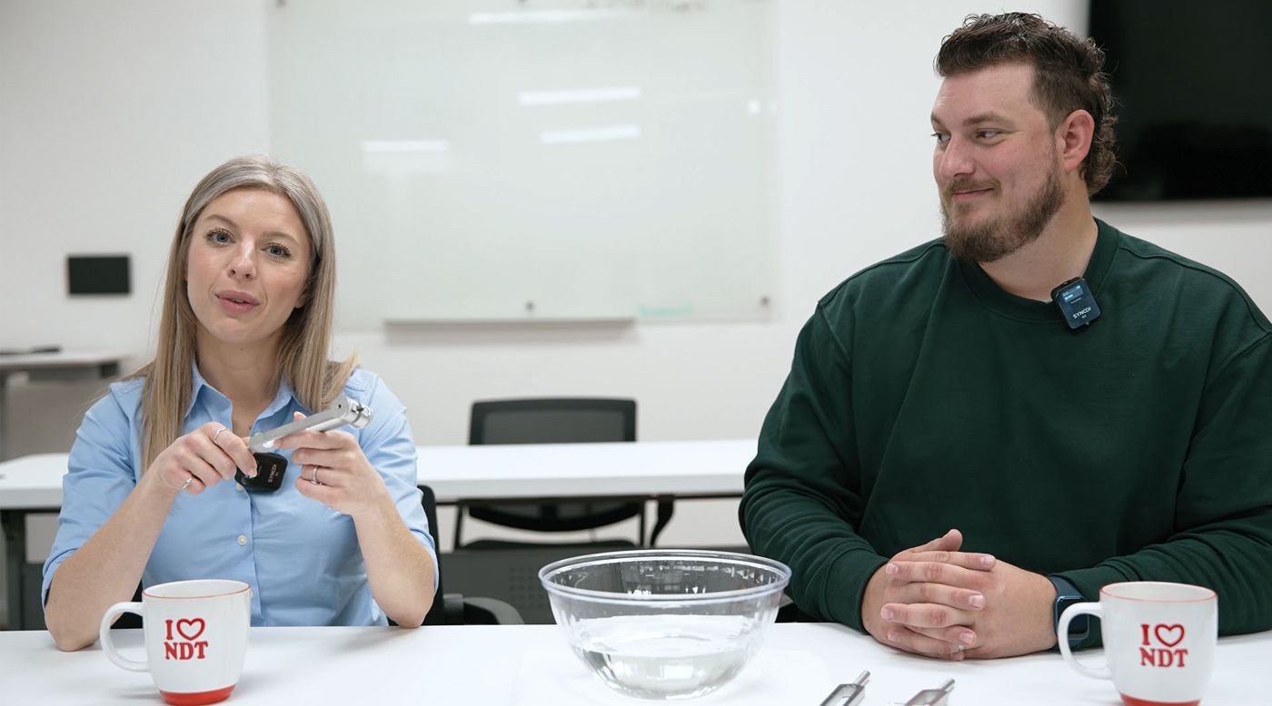 Two people sit at a table with a clear bowl of water. The woman holds a tuning fork, and both appear engaged in conversation.