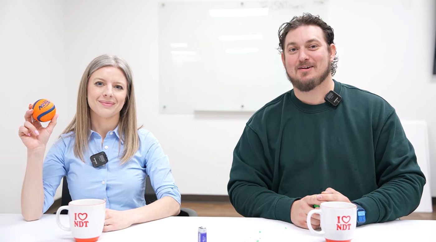 Two people sitting at a table with "I ♥ NDT" mugs. The woman holds a small basketball. Both wear clip-on microphones.