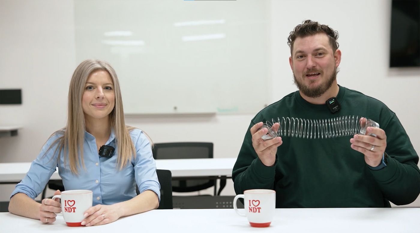 Two people sit at a table with mugs. The man holds a metal slinky. A whiteboard is visible in the background.