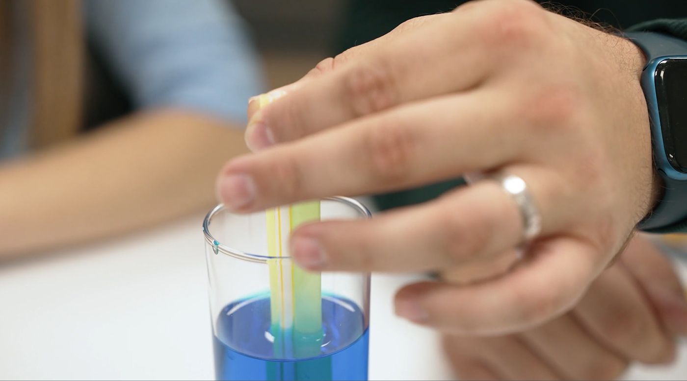 A hand holding a pipette above a beaker filled with blue liquid, with a person in the background.