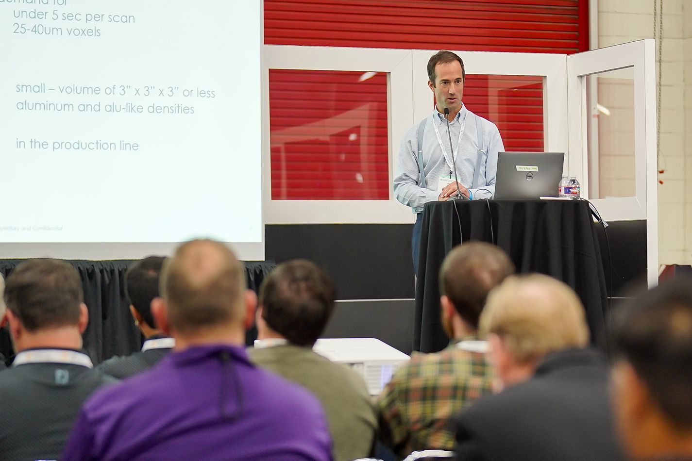 A speaker presenting to a small audience, with a screen displaying text in the background.