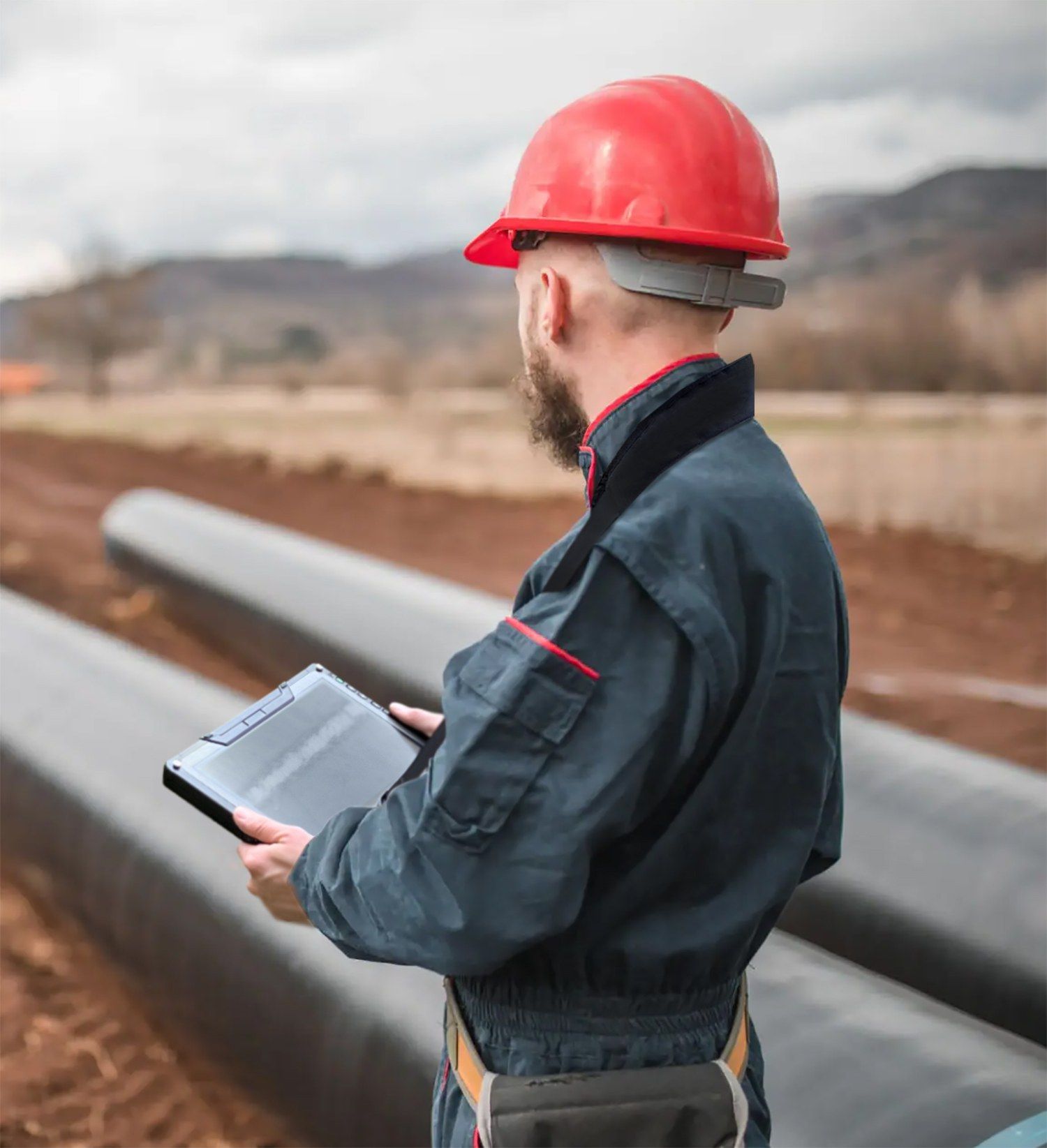 Inspector in a red hard hat reviewing a tablet beside pipeline sections in the field