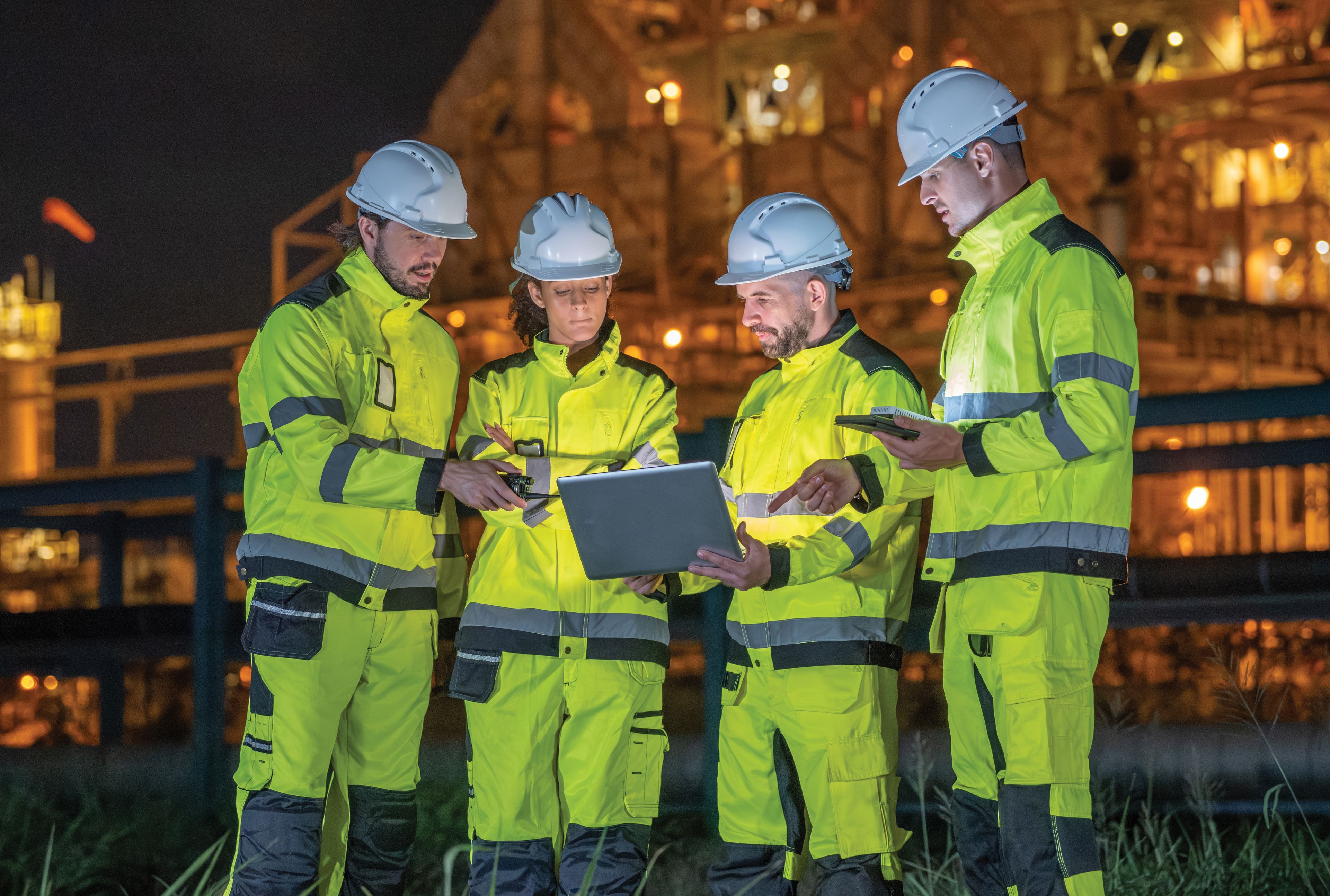 A team of four workers in hi-vis gear and hard hats reviews a laptop at night at an industrial facility.