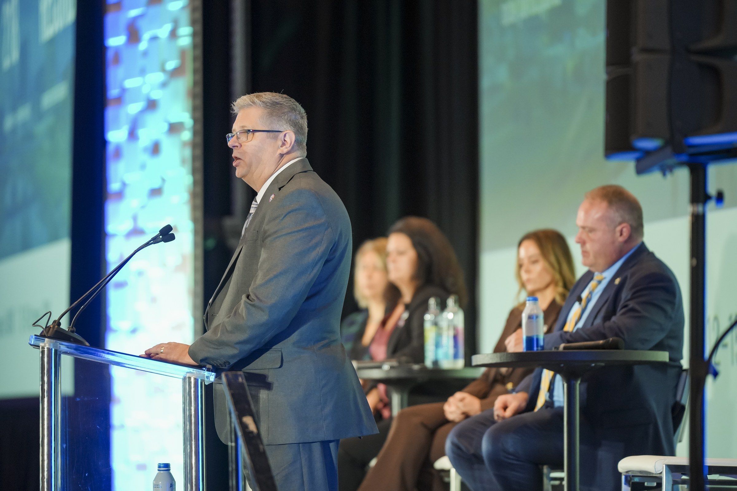 ASNT Interim CEO Barry Schieferstein speaking at a podium with ASNT Board members seated onstage behind him