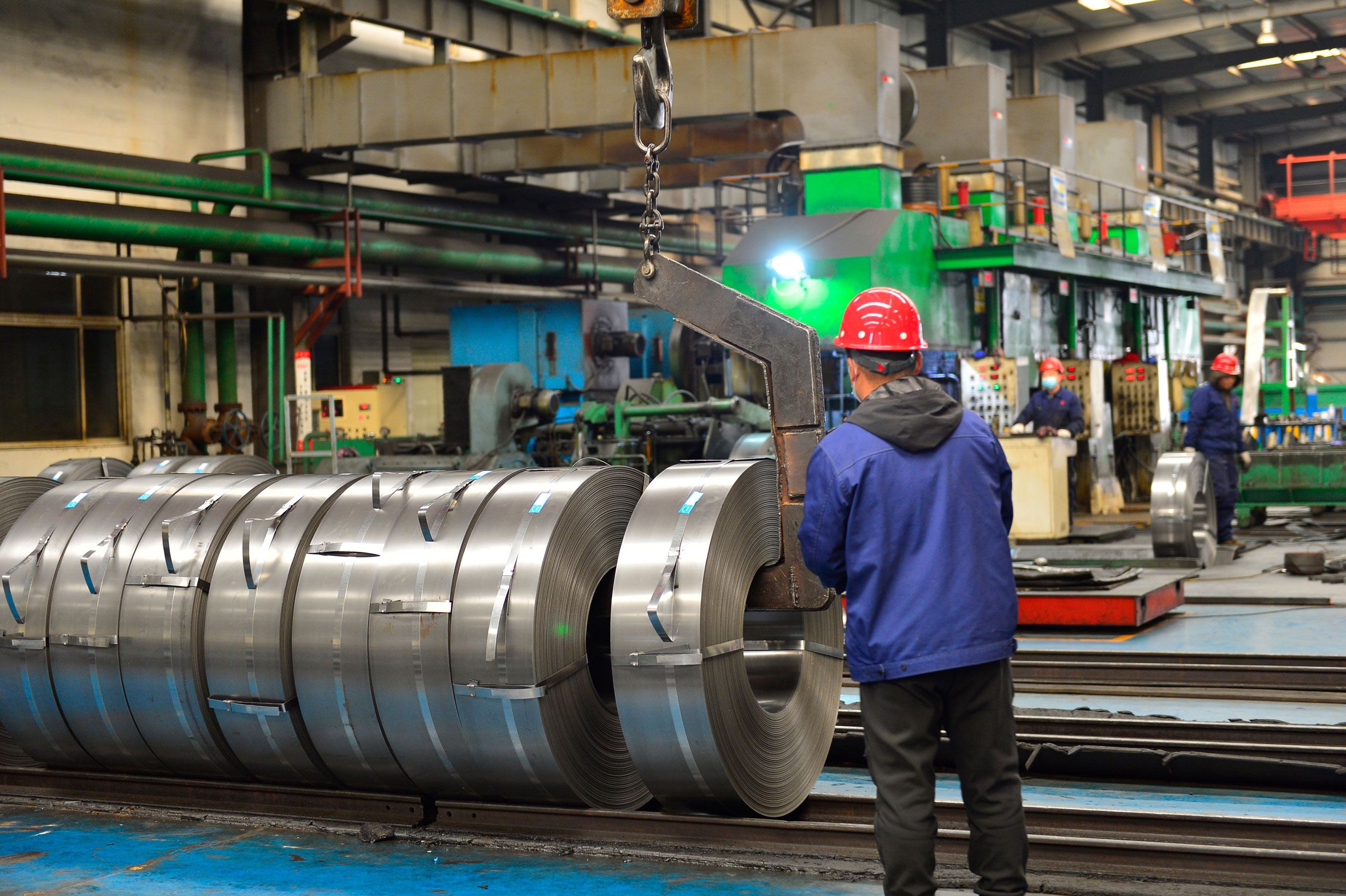 Worker in a red hard hat and blue jacket beside a row of steel strip coils being lifted by a crane hook in a steel mill