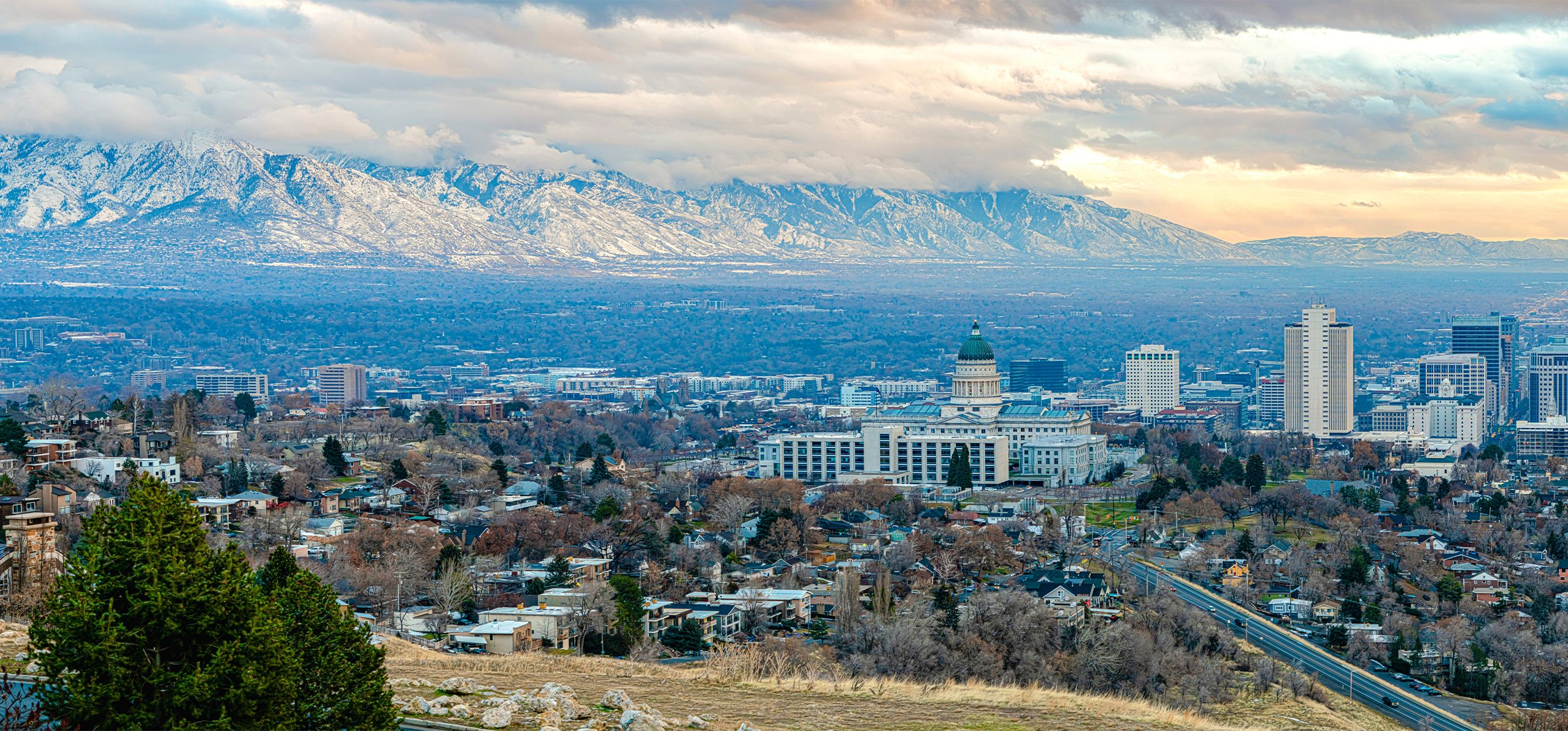 Aerial view of Salt Lake City with the Utah State Capitol, surrounded by urban buildings and snow-capped mountains under a cloudy sky.