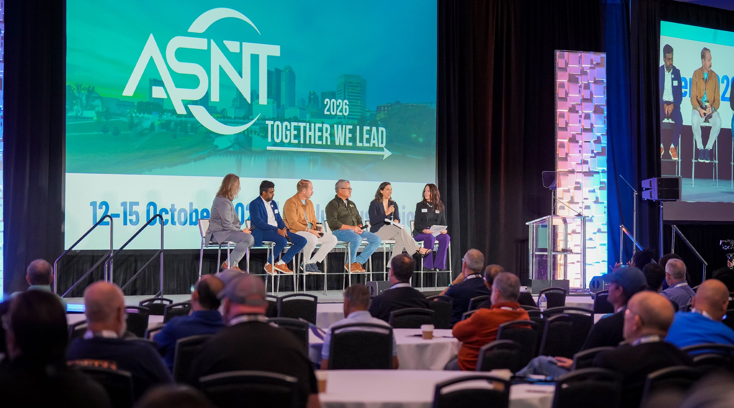 A panel discussion at the ASNT event with six speakers seated on stage, audience members watching attentively.
