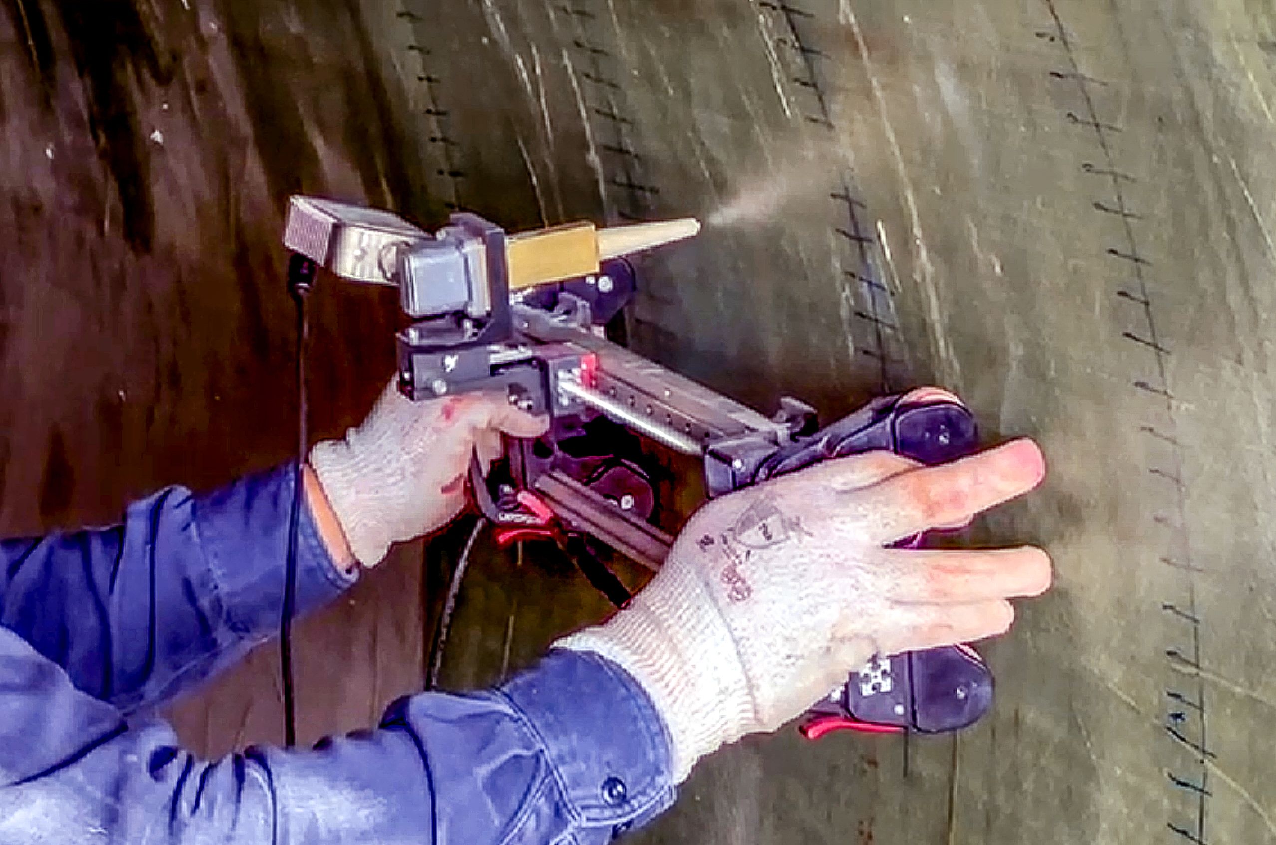 A person operates a handheld microwave testing device on a large pipe surface, wearing protective gloves and a jacket.
