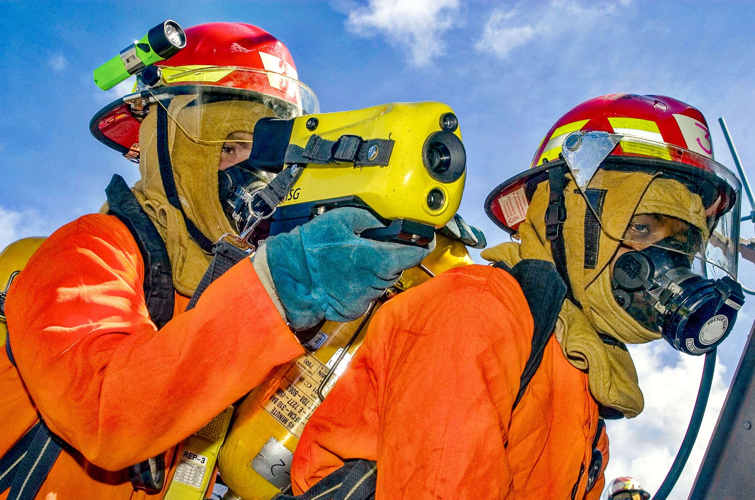Firefighters in orange gear and red helmets use a thermal imaging camera during a drill, set against a clear blue sky.
