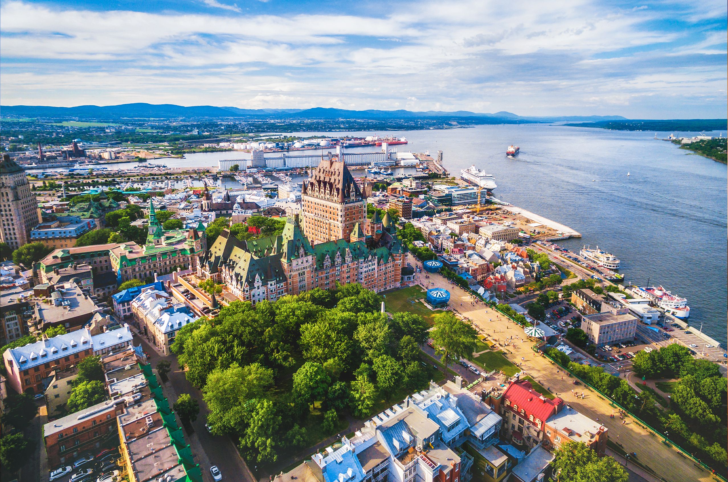 Aerial view of Québec City featuring the historic Château Frontenac, surrounded by colorful buildings, greenery, and a river with ships.