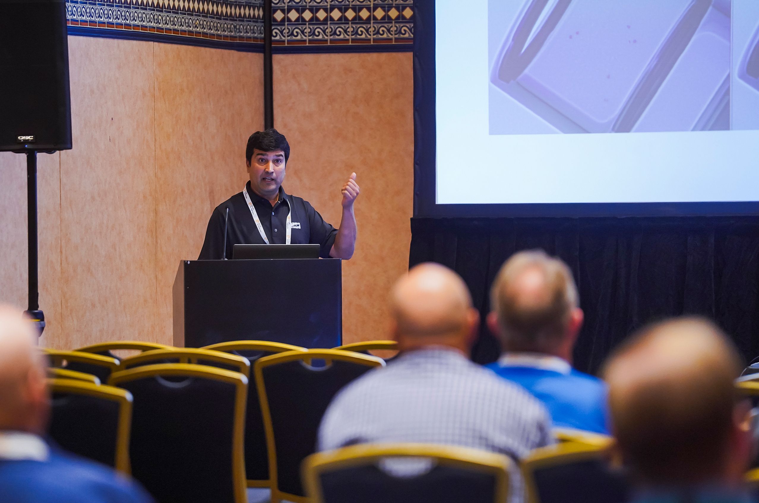 A man giving a presentation to an audience in a conference room, gesturing with one hand near a screen displaying a slide.