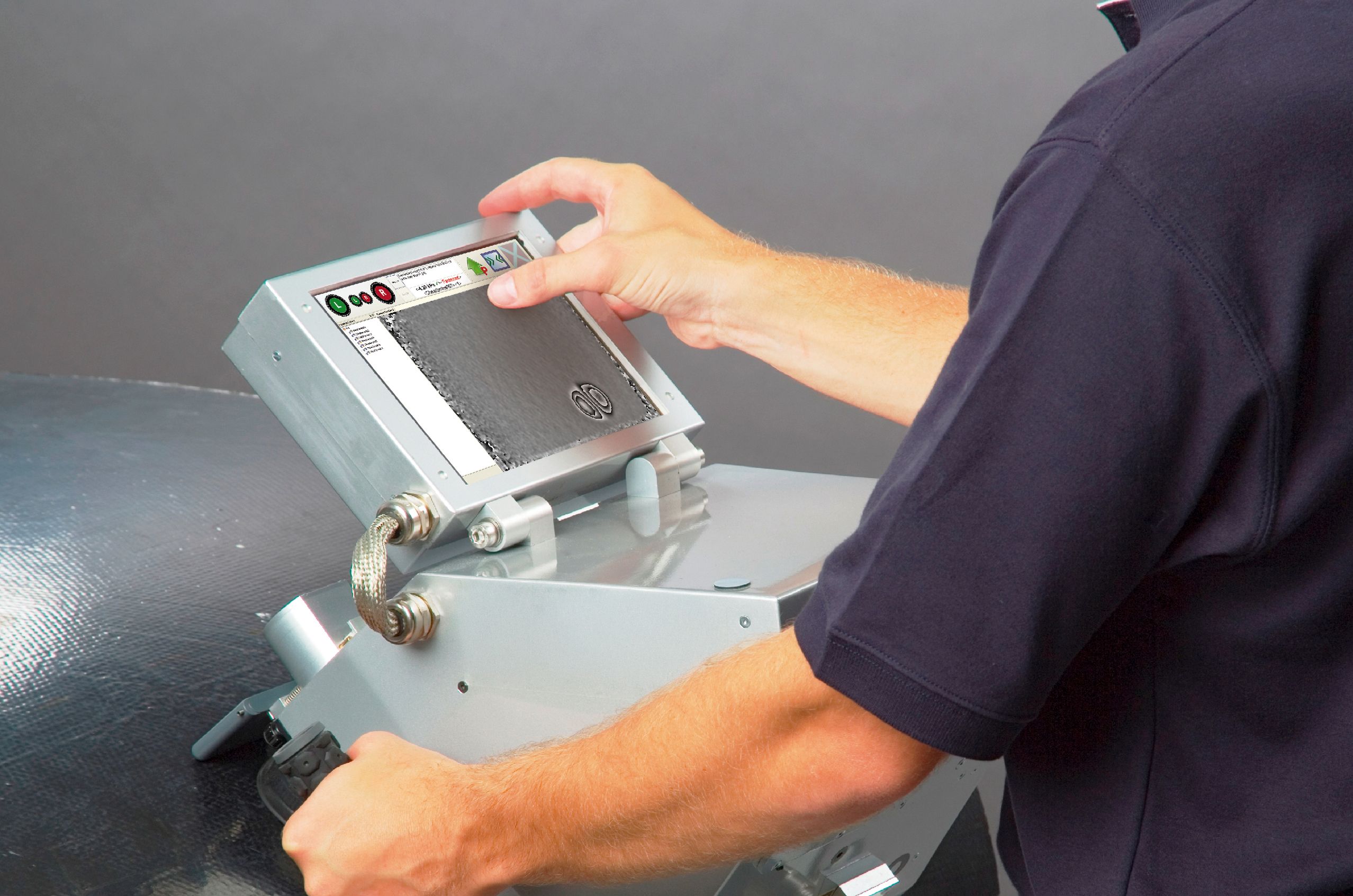 Person operating a handheld metal shareography device with a digital screen, adjusting settings, against a textured black surface.