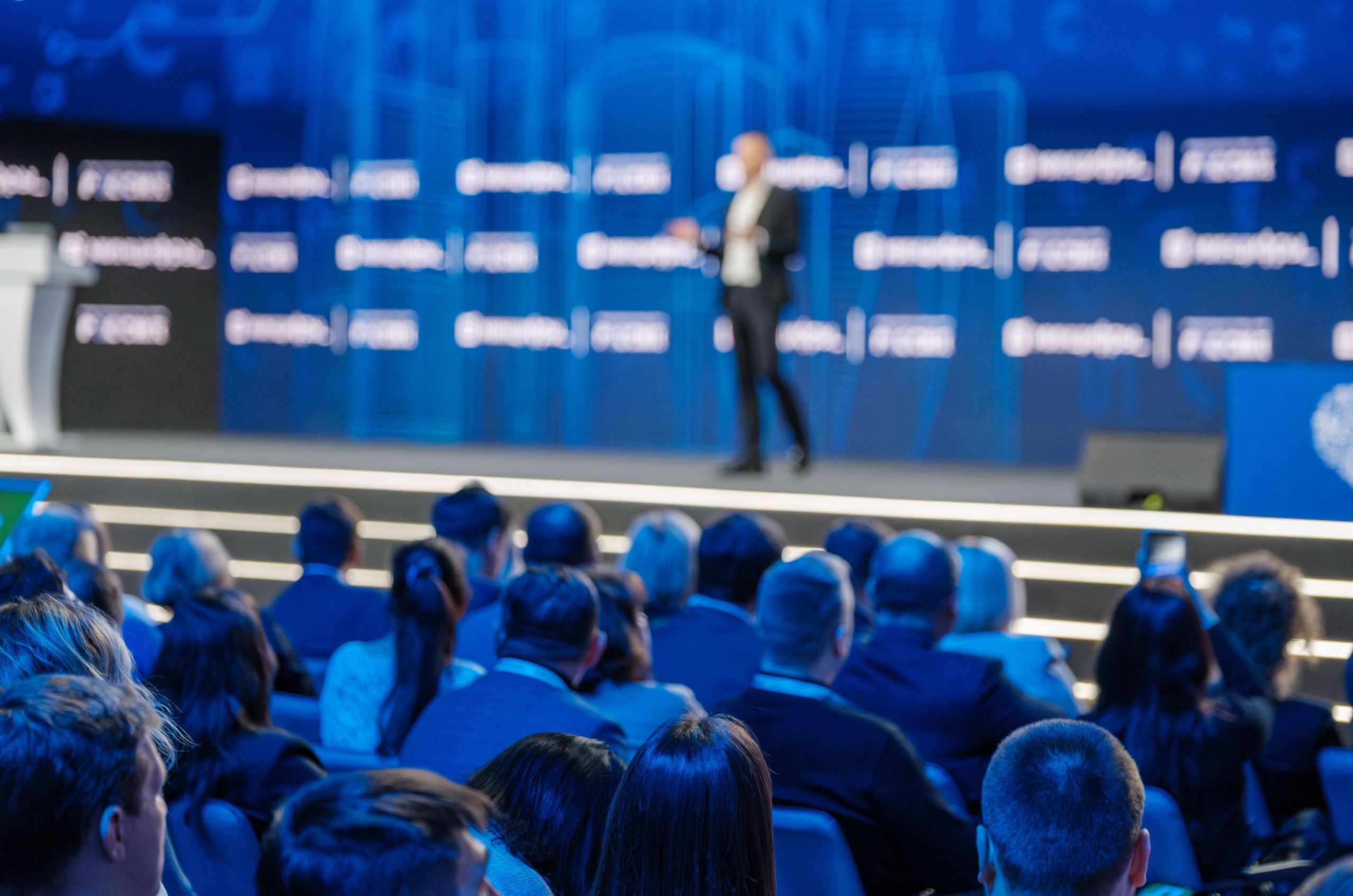 Blurred speaker on stage at a conference with a seated audience watching, blue-lit background featuring event branding.