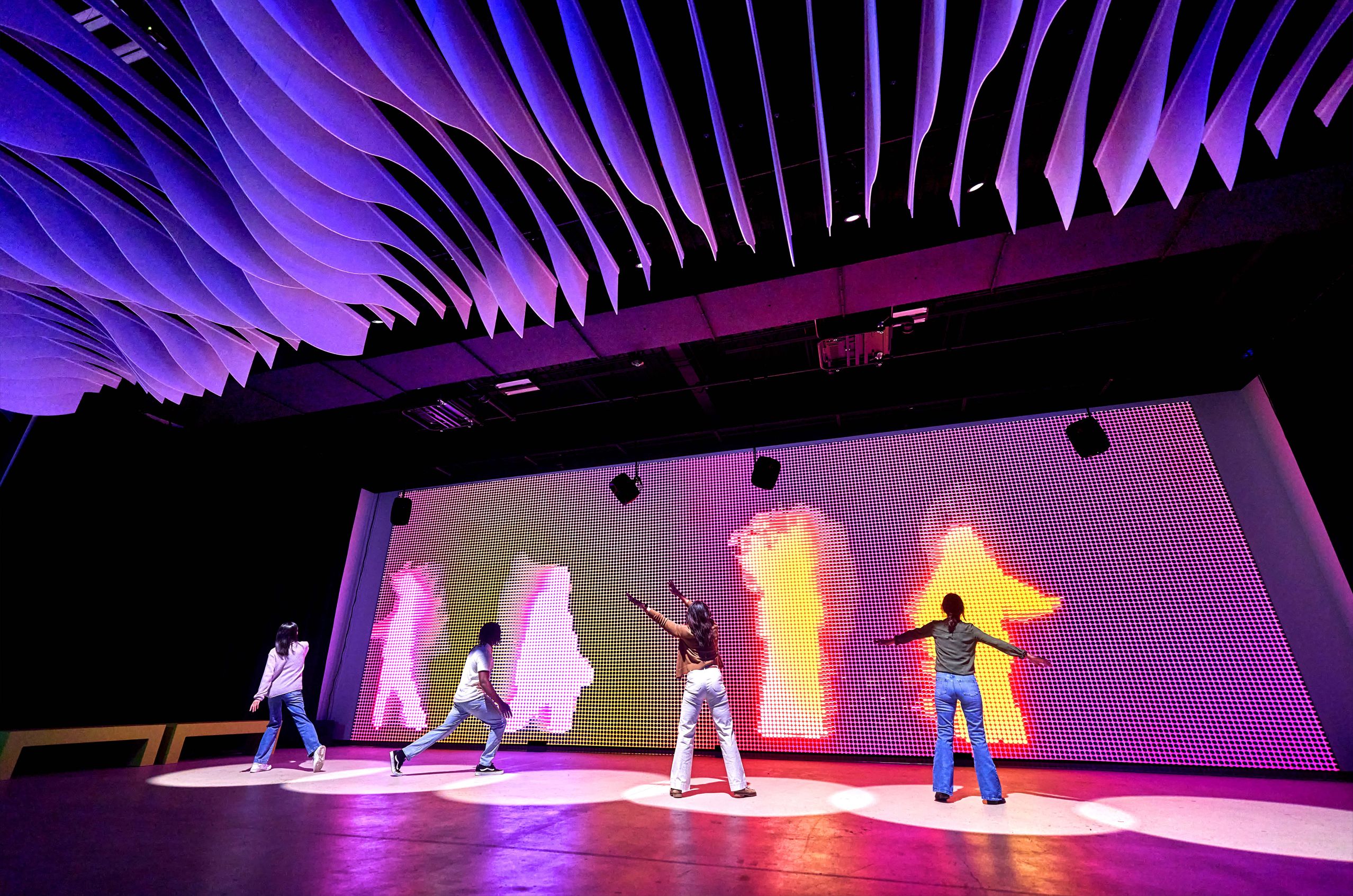 Four people dance in front of a vibrant digital screen with colorful silhouettes under a ceiling with wavy panels.