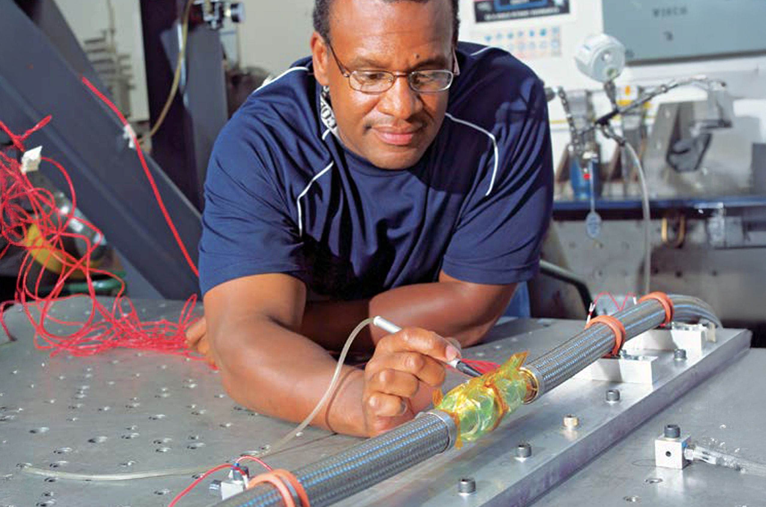 Technician inspects a metallic setup with various cables and components in an industrial setting.