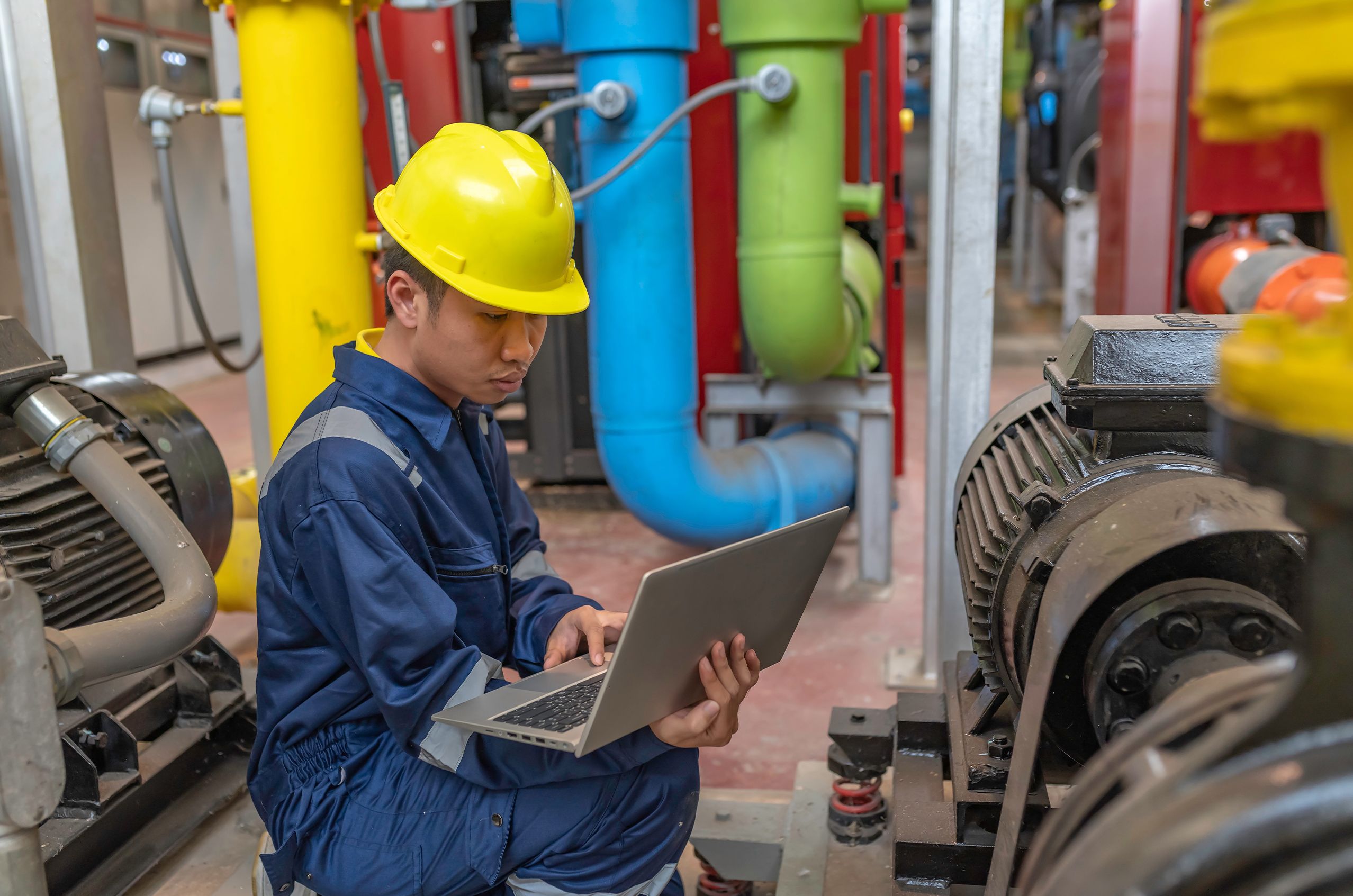 NDT worker in a hard hat and overalls uses a laptop in an industrial setting with colorful pipes and machinery in the background.