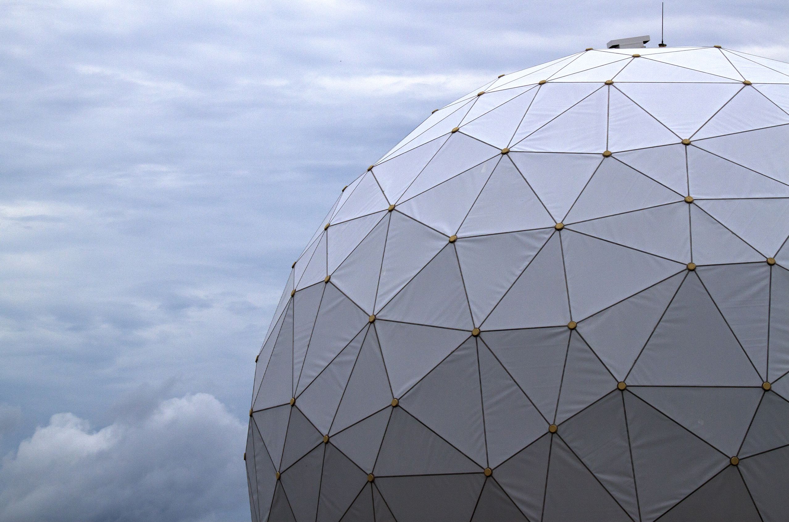 A radome, a geodesic dome with triangular panels, against a cloudy sky background, featuring a white surface and gold nodes.