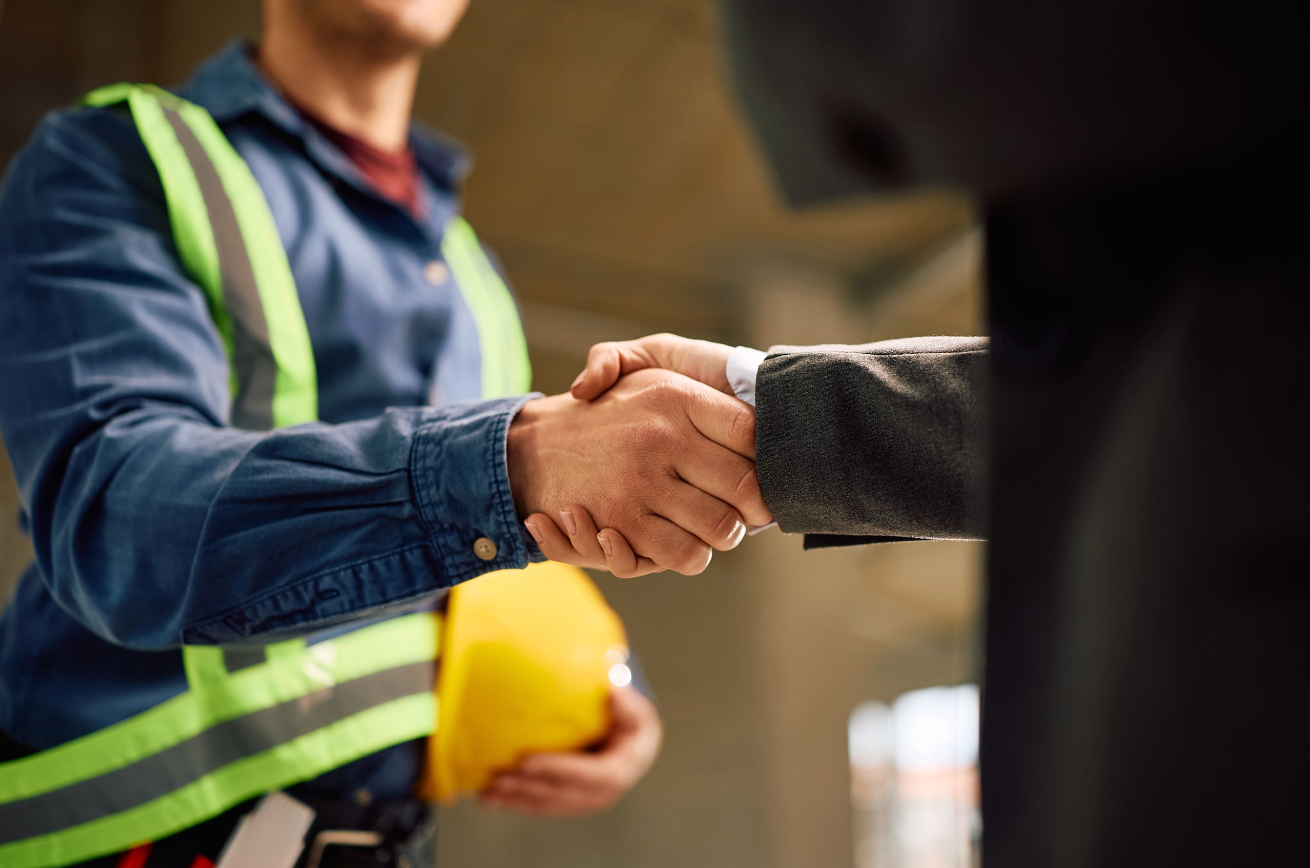 A NDT technician in a reflective vest shakes hands with a person in a suit, holding a yellow hard hat in the background.
