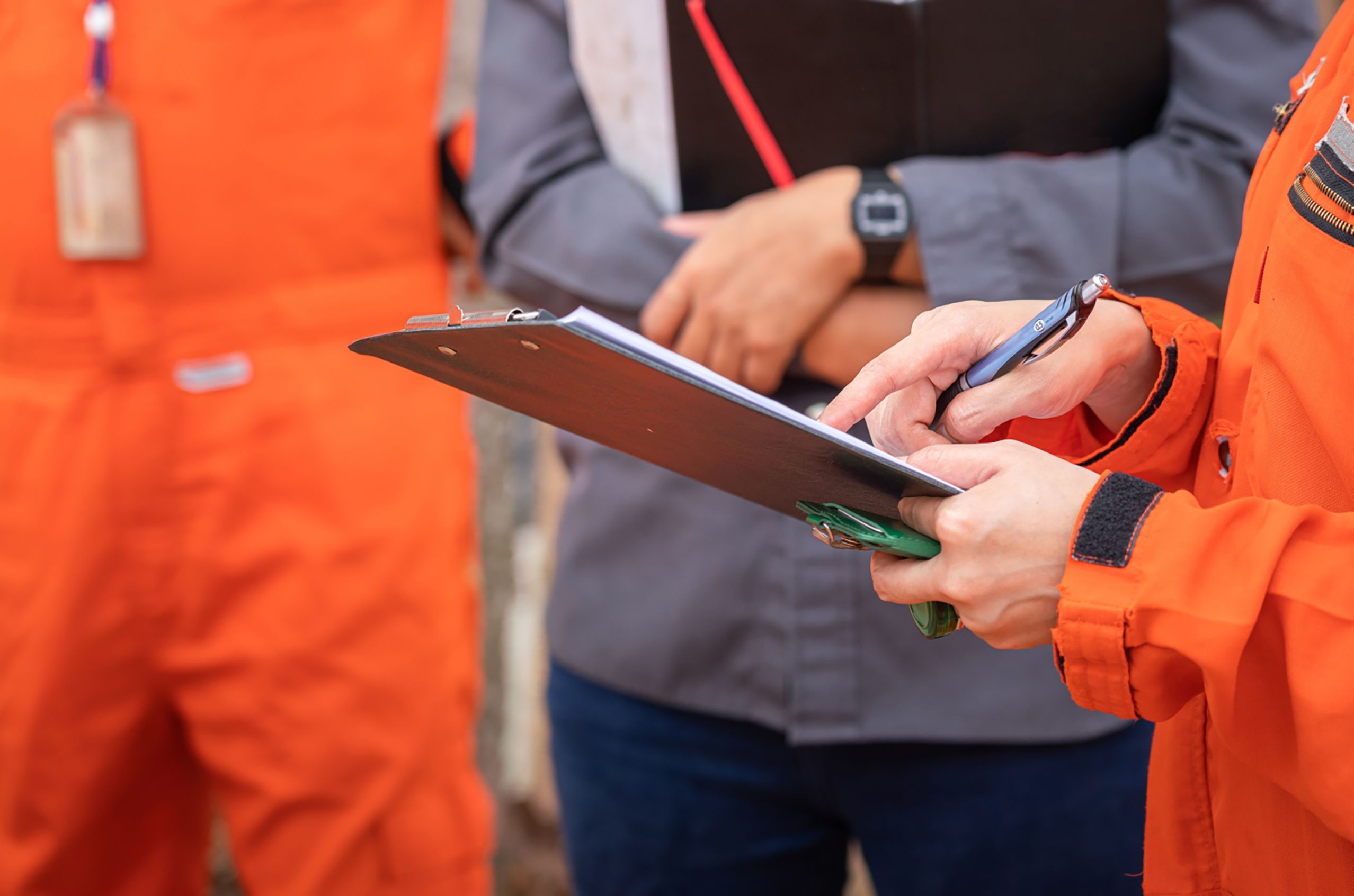 Close-up of a person in an orange safety uniform holding a clipboard and a pen, writing notes. Two other people stand in the background with arms crossed.