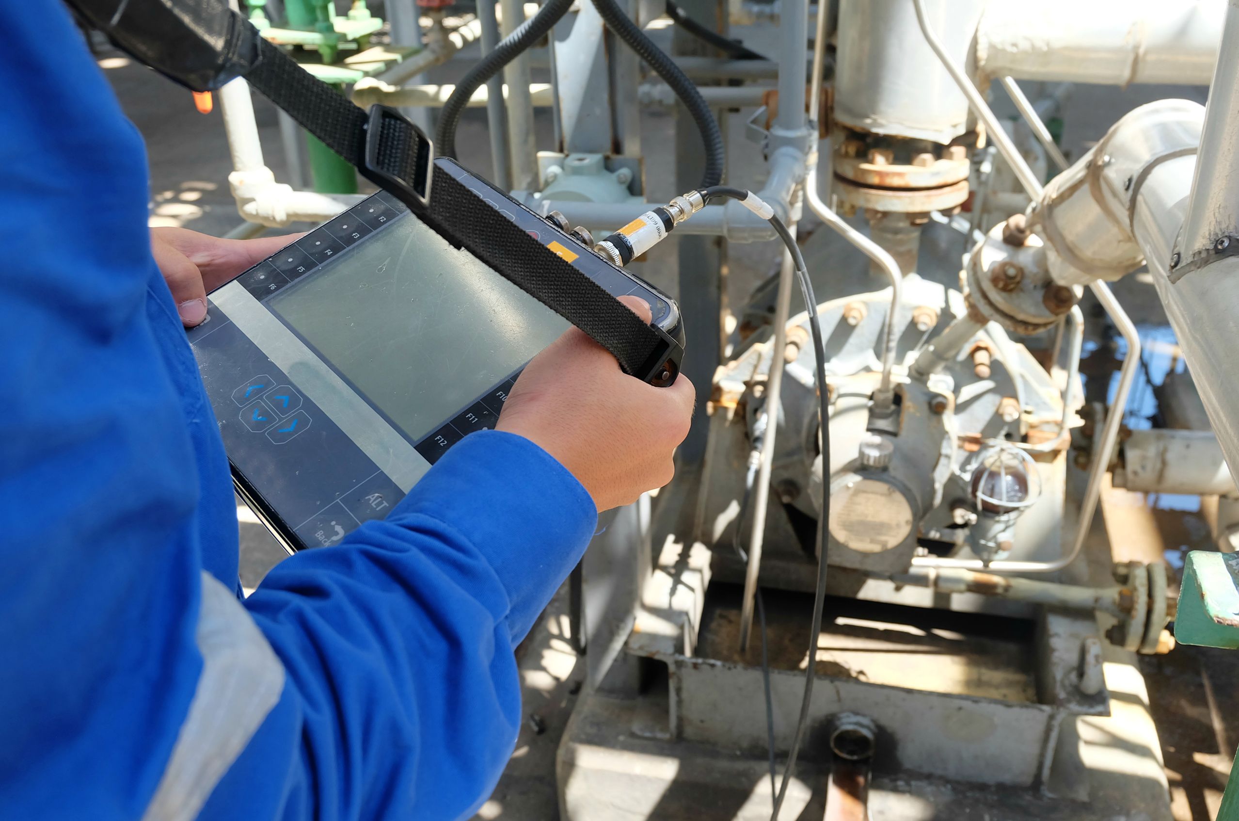Person in blue uniform uses a handheld device to inspect industrial machinery with pipes and gauges.