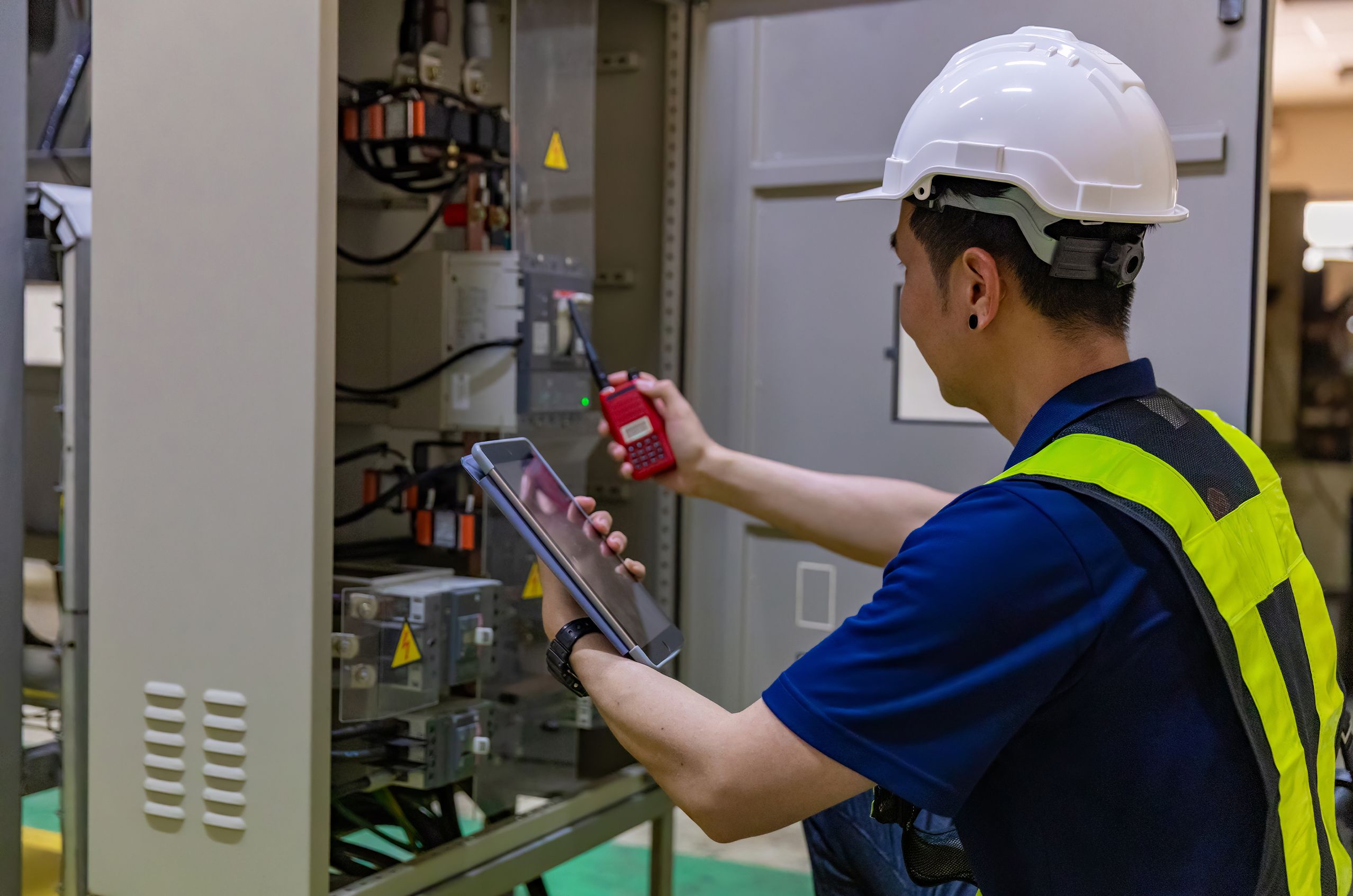 Electrician in a hard hat and safety vest checks a control panel, using a tablet and radio for communications.