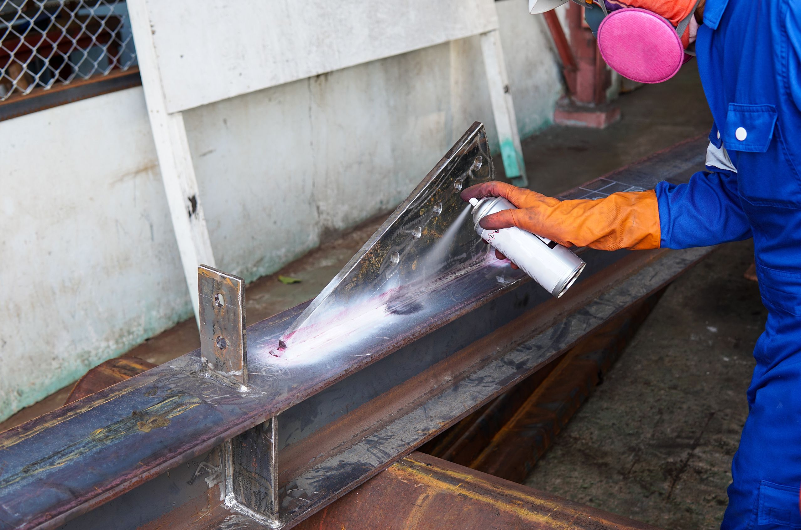 A technician wearing safety gear sprays a penetrant on a metal beam during a dye penetrant inspection in an industrial setting.