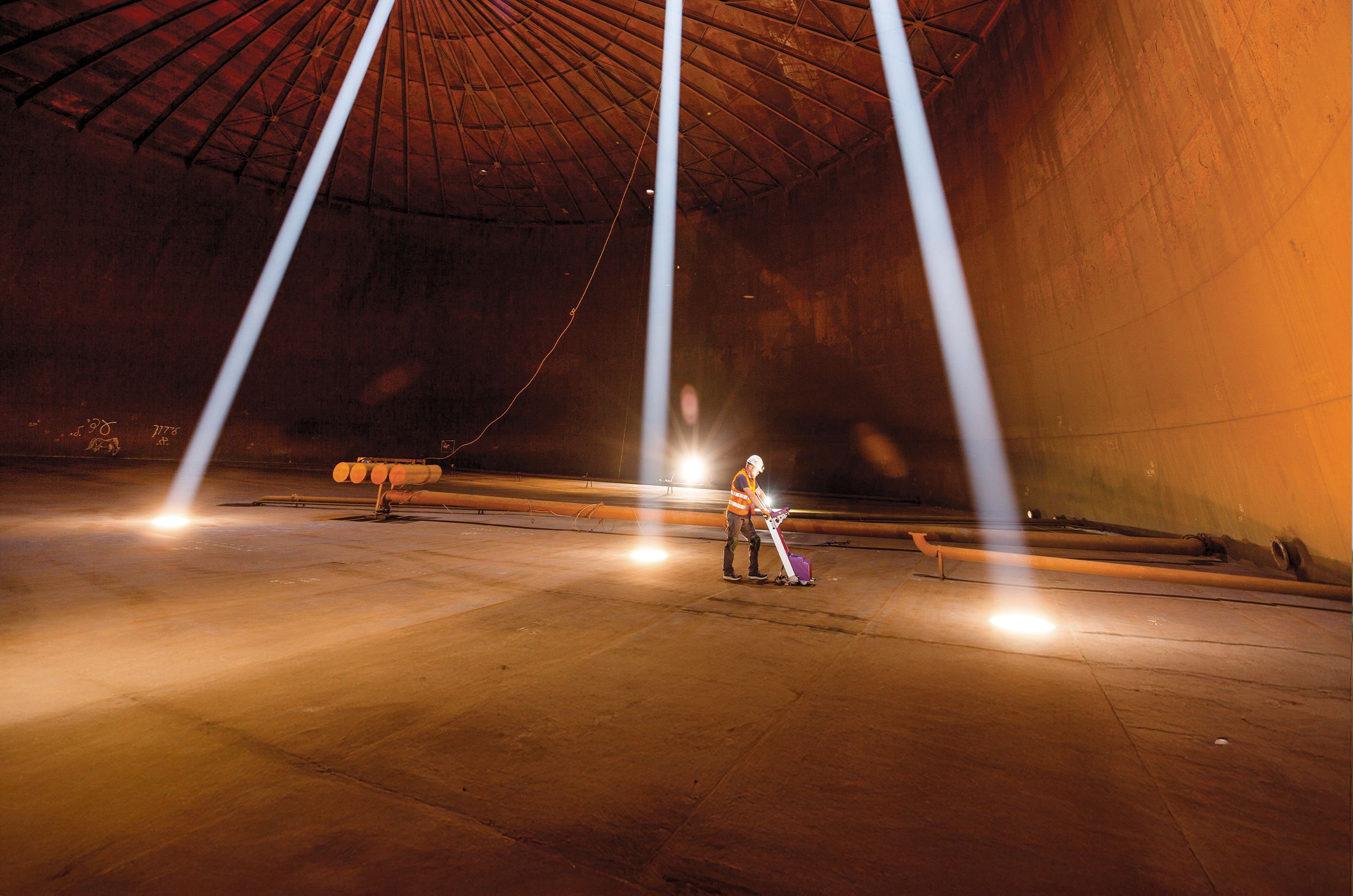 Person in safety gear using equipment inside a large industrial tank, illuminated by beams of light from above.