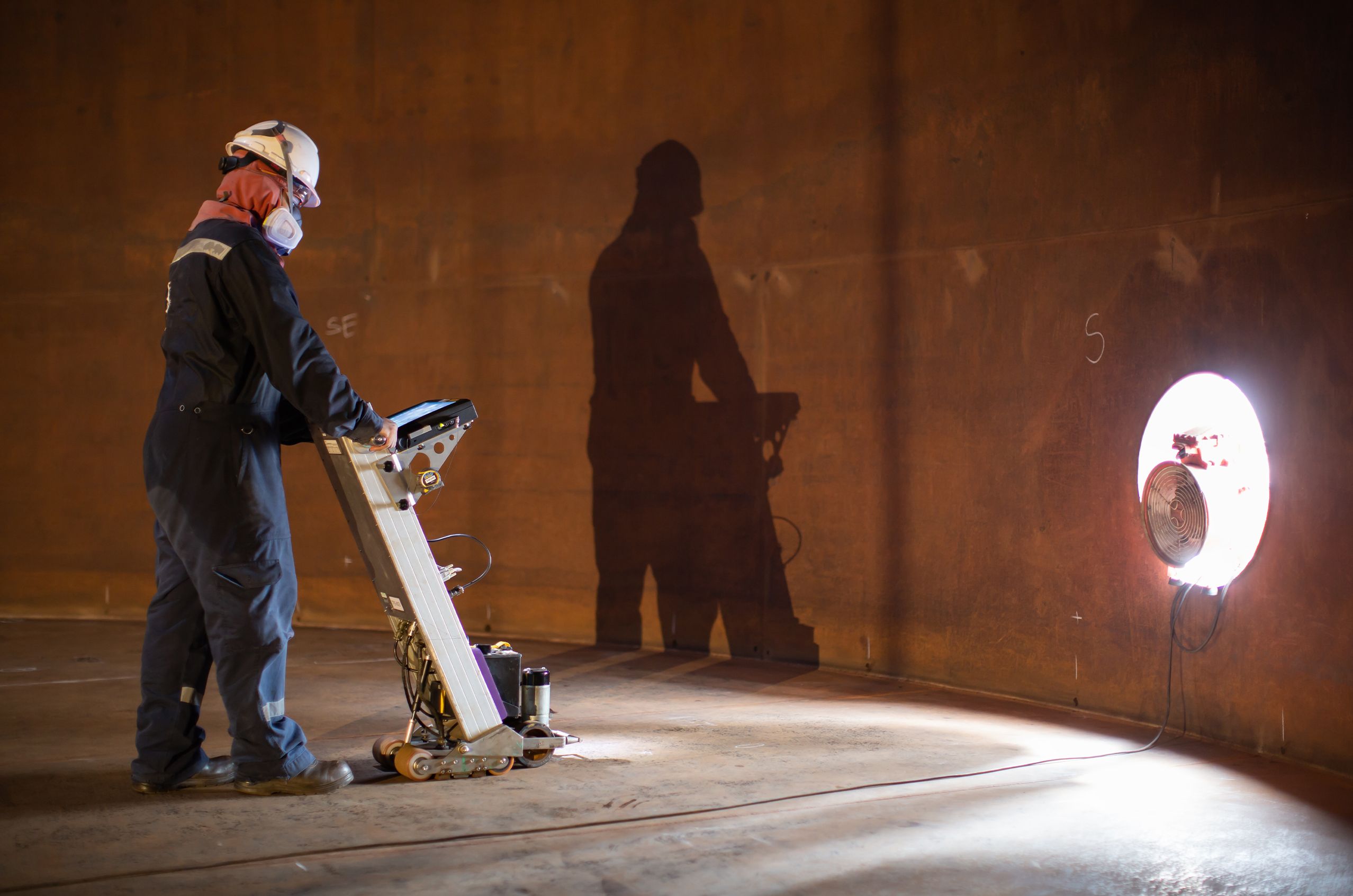 A worker in protective gear operates a machine inside a dimly lit tank, casting a shadow on the wall near a bright light source.