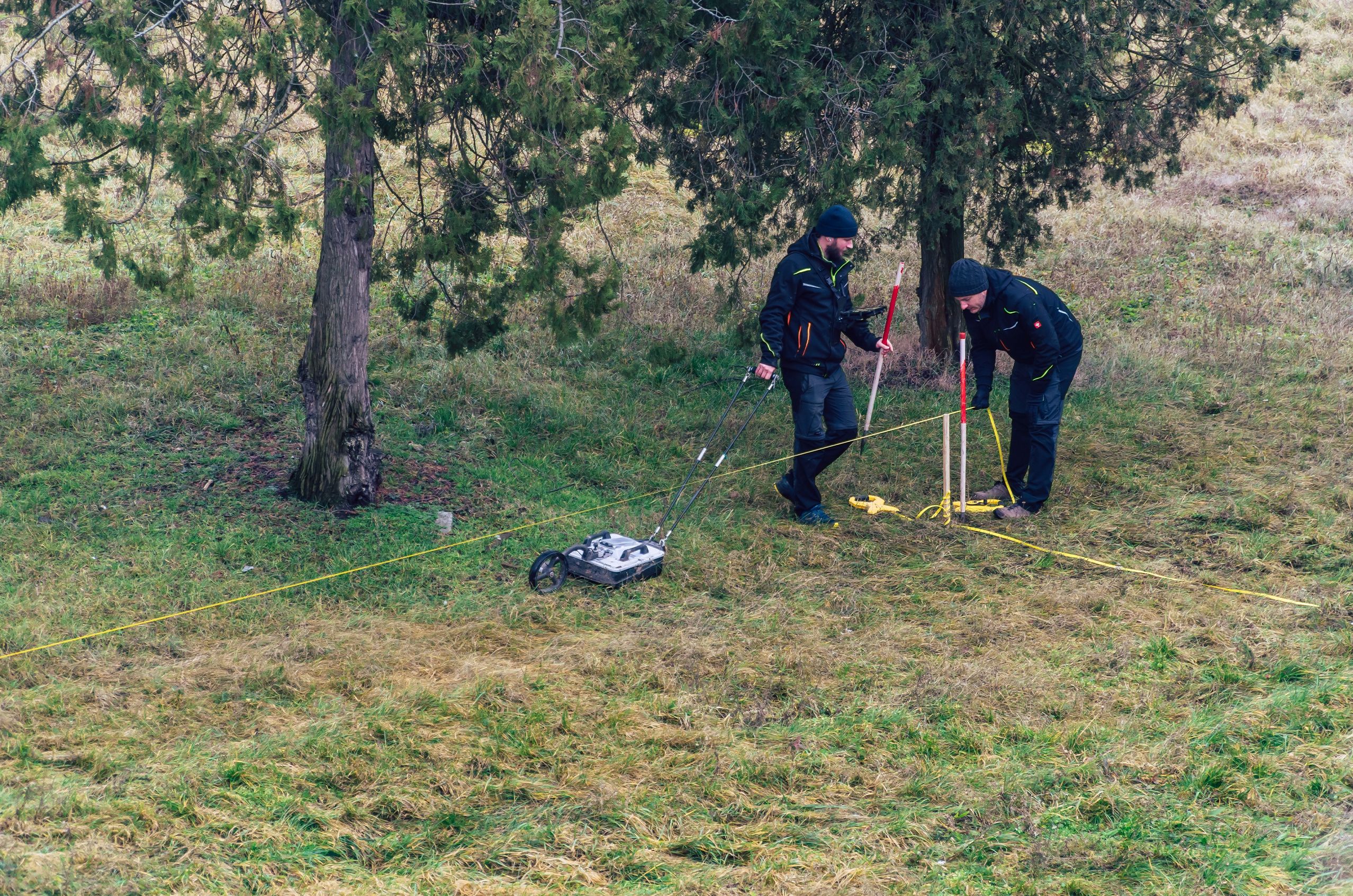 Two people in dark clothing use surveying equipment on a grassy field under trees, with measuring tape and a wheeled instrument.