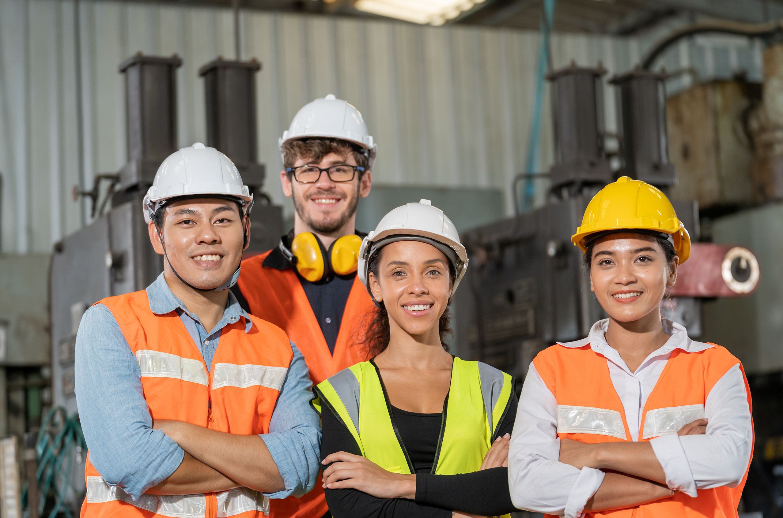 A diverse team of industrial workers wearing safety helmets and uniforms, standing in a line with arms crossed. The focus is on a smiling woman at the front, with her colleagues blurred in the background, suggesting unity and teamwork in an industrial setting.