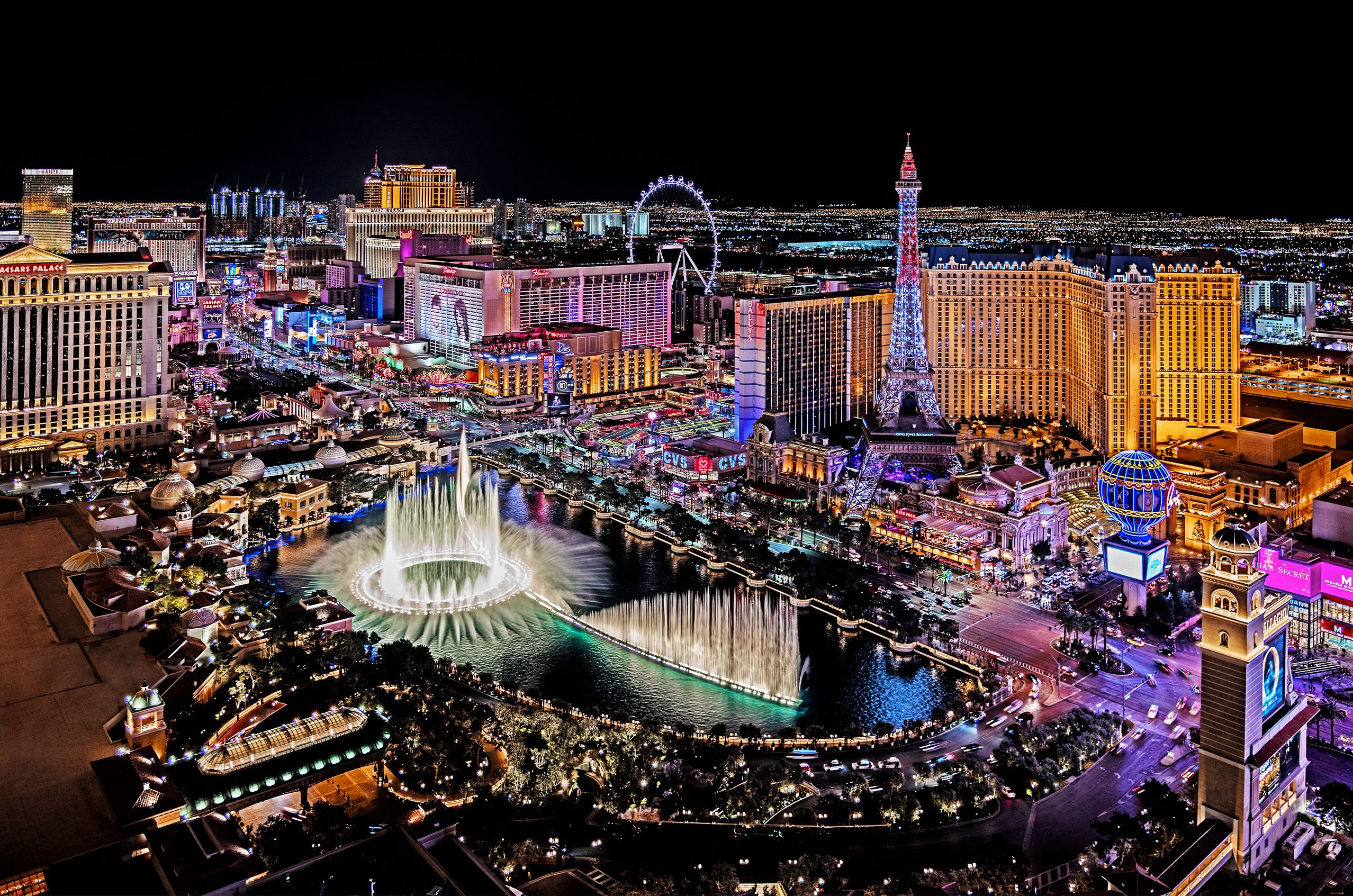 Nighttime aerial view of the Las Vegas Strip, brightly lit with colorful lights and featuring iconic landmarks such as the Bellagio fountains, the Eiffel Tower replica, and various hotels and casinos.