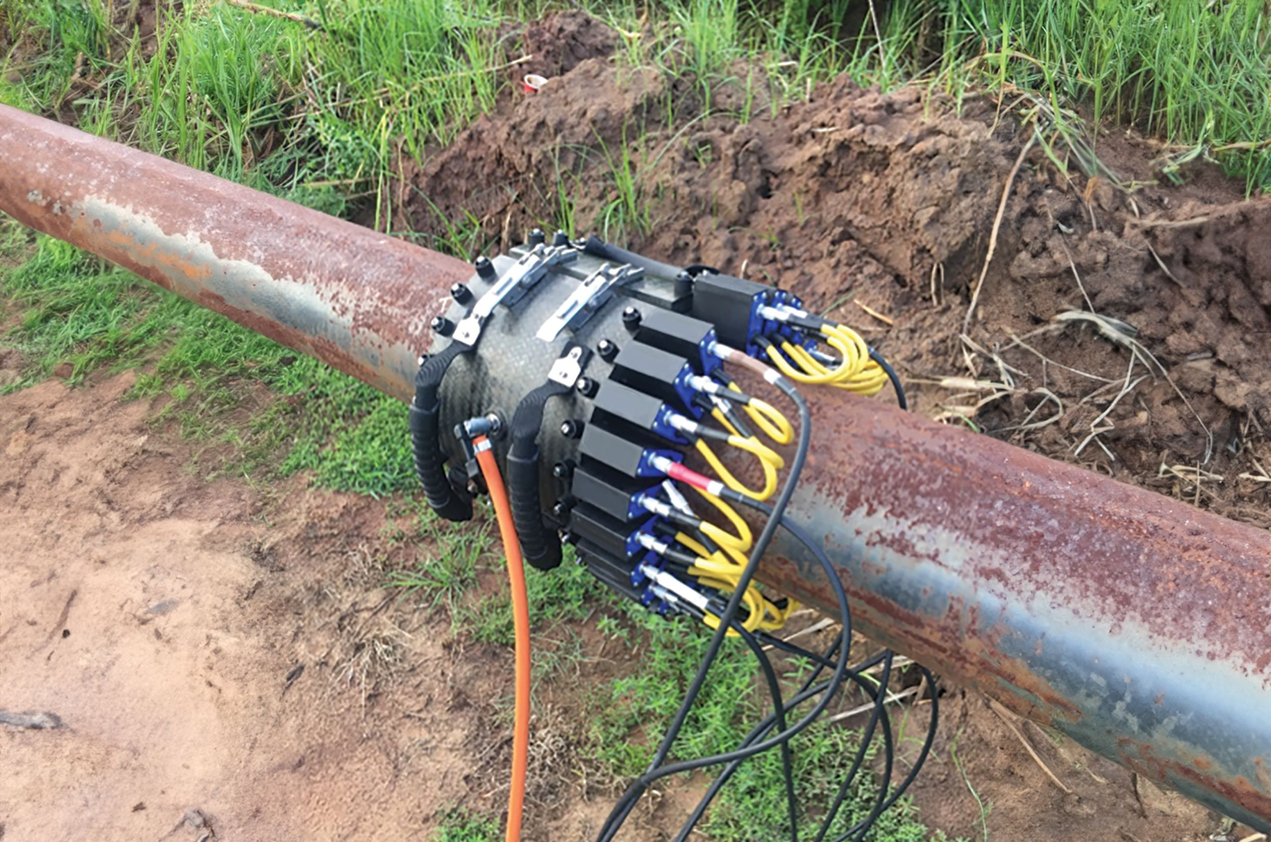 A guided wave testing device is attached around a rusty pipeline outdoors. The device features multiple sensors and cables wrapped around the pipe to inspect for defects or corrosion. The surrounding area is grassy with exposed soil and vegetation.